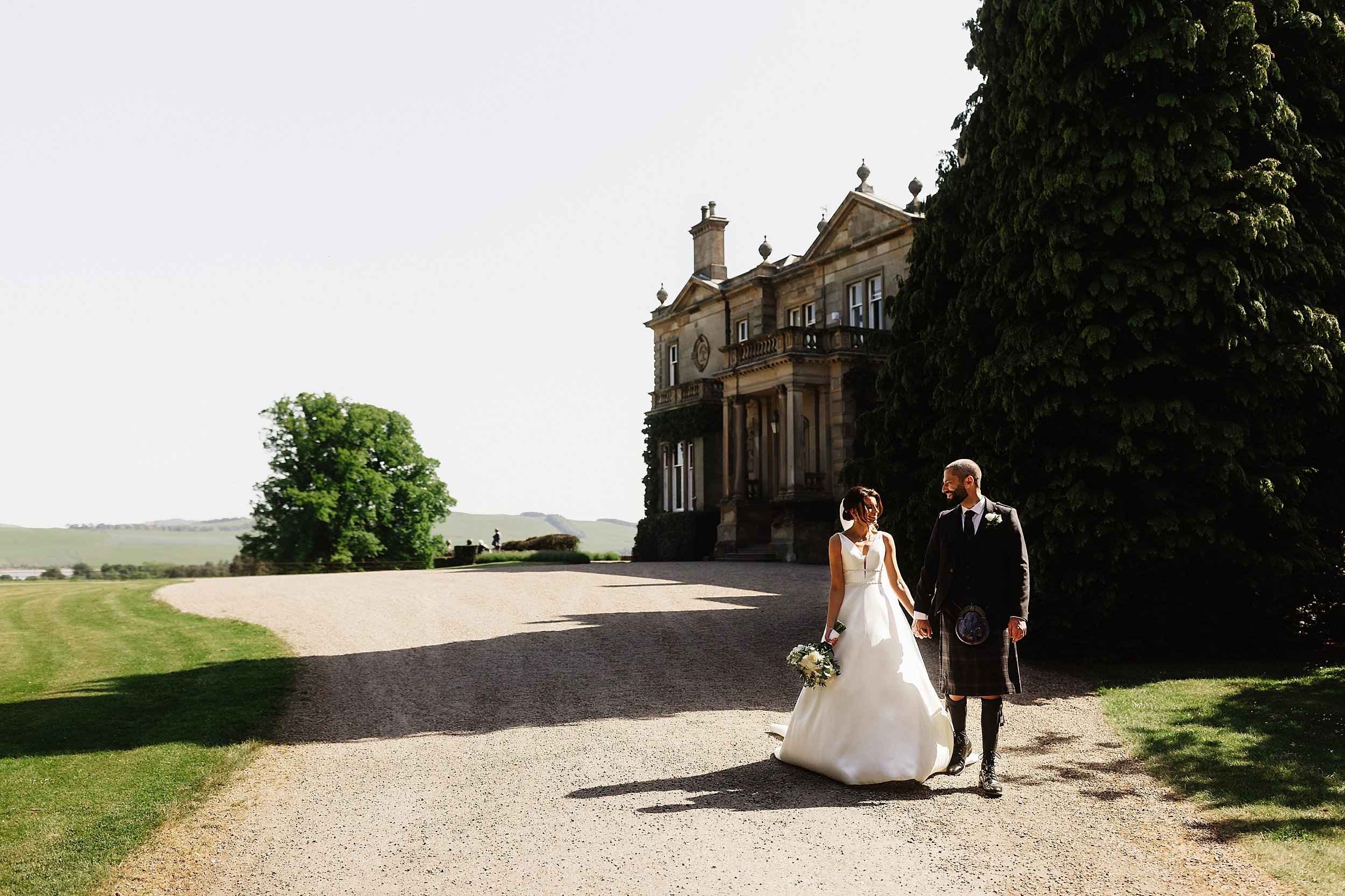 the bride and groom walk along a path away from errol park wedding venue the house trees and hills are visible in the background