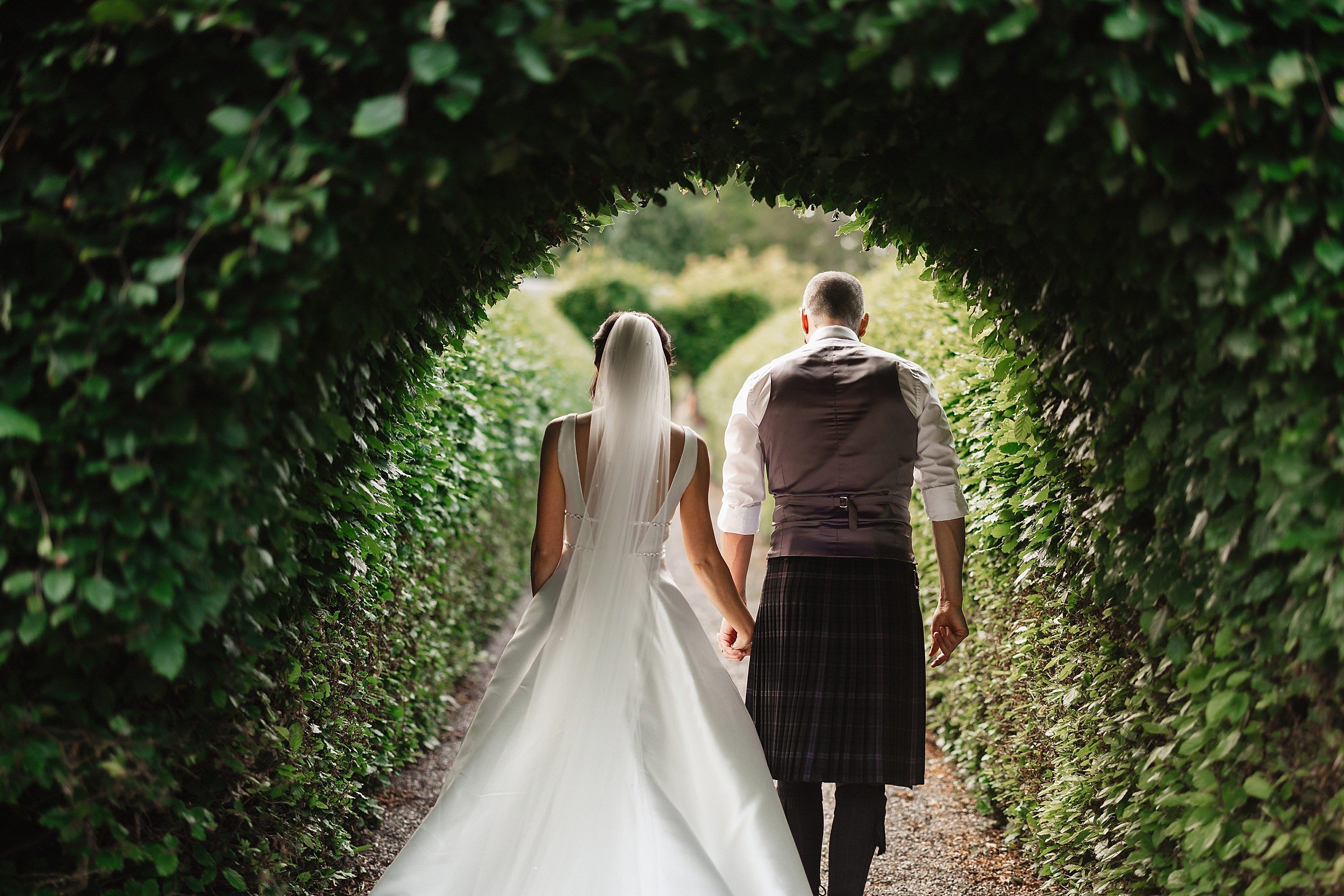 the bride and groom walk away hand in hand through a topiary arch along a path lined with high green hedges in the gardens of errol park wedding venue in perthshire in scotland