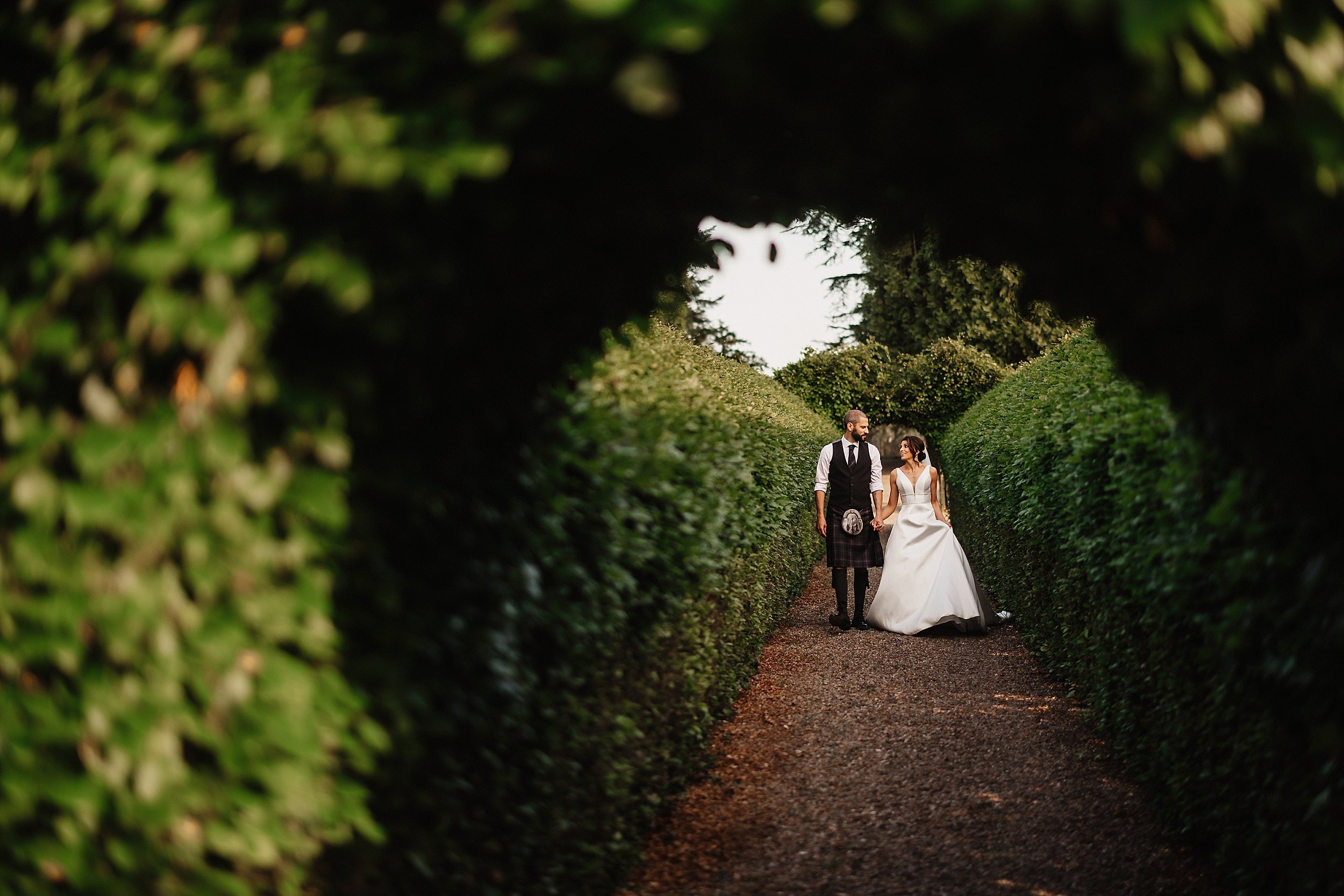 the bride and groom walk hand in hand along a path lined with high green hedges in the gardens of errol park wedding venue in perthshire in scotland