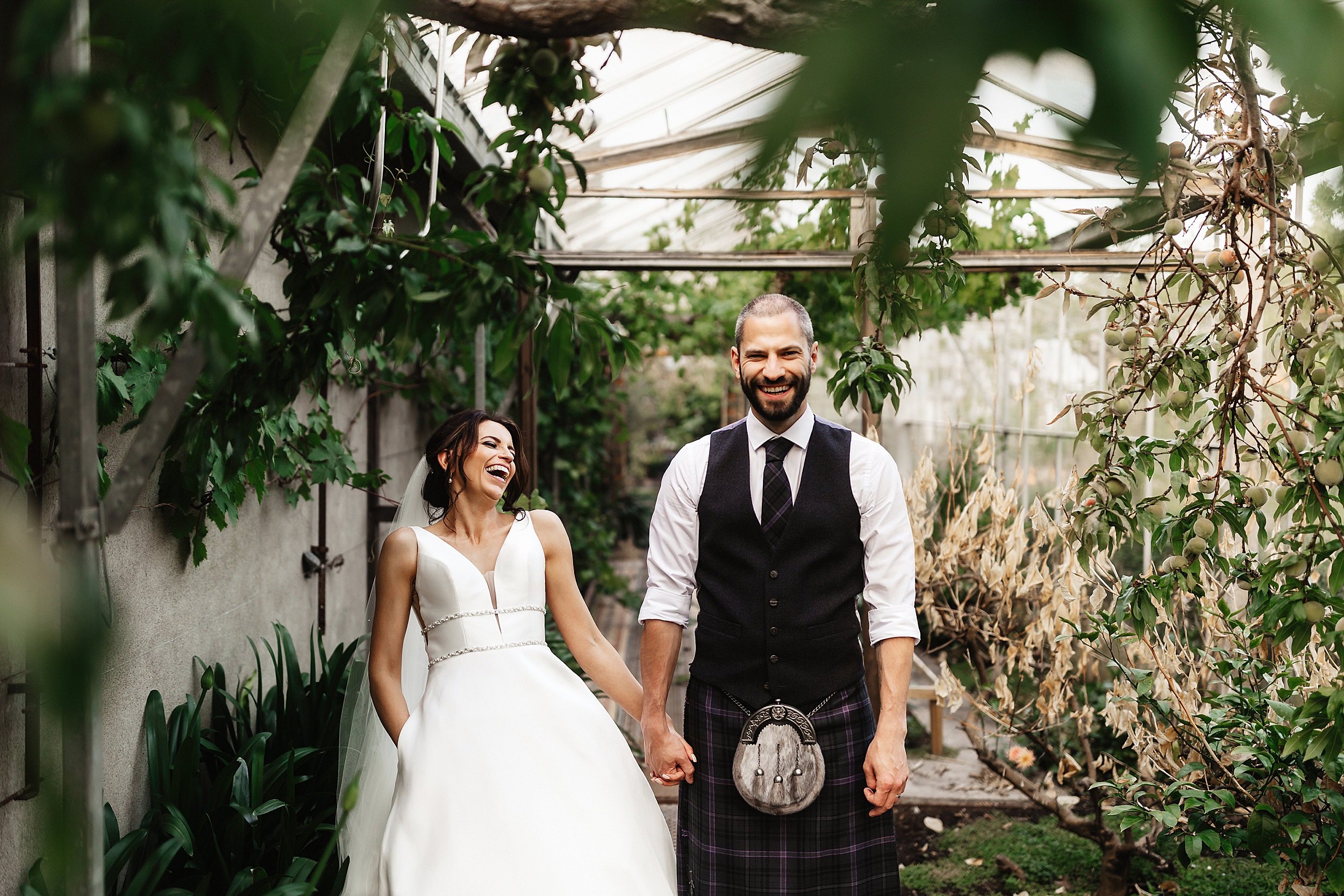 the bride laughs as she stands hand in hand with the groom in a greenhouse filled with plants in errol park wedding venue in perthshire in scotland