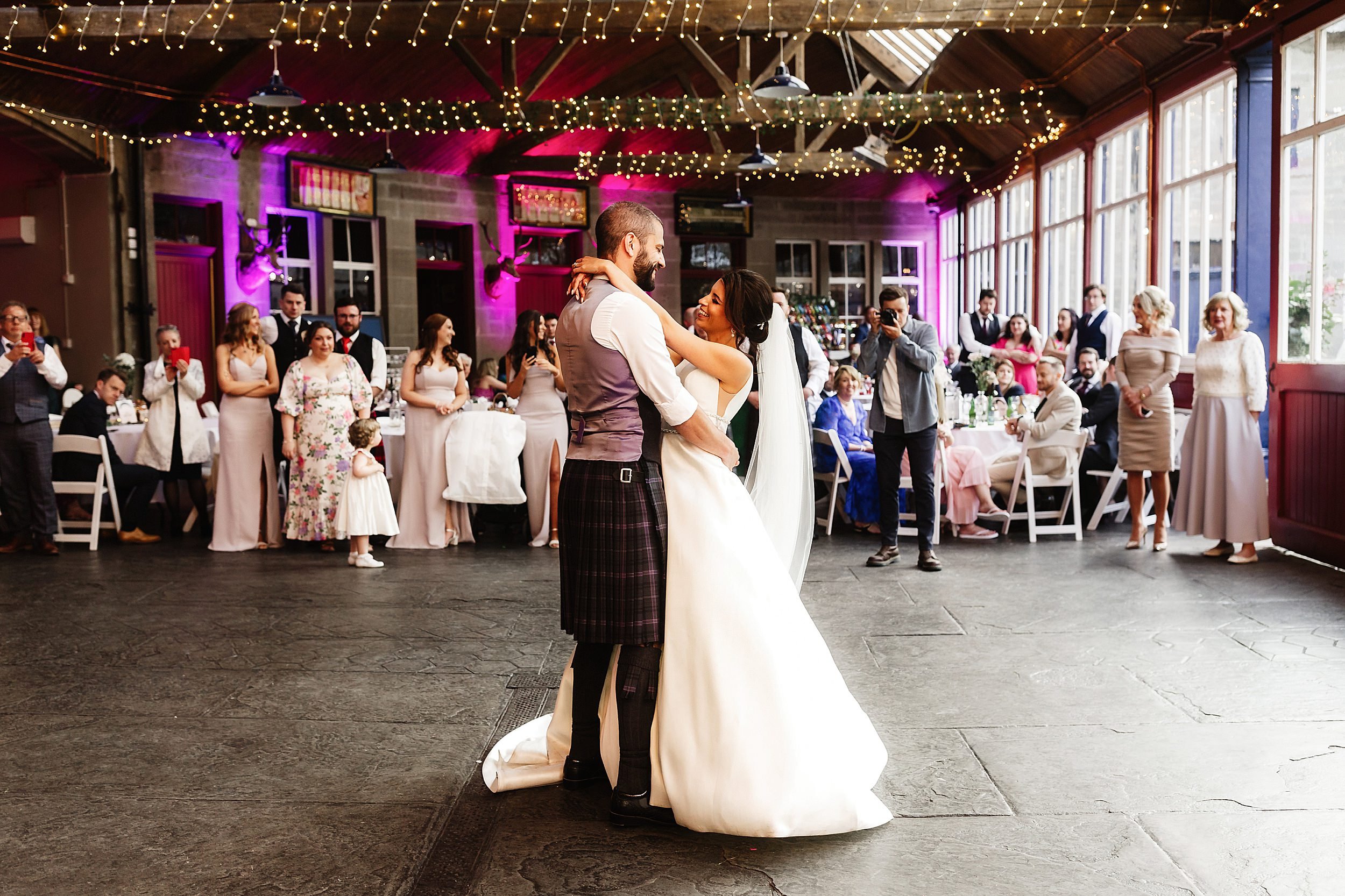 guests look on as the bride and groom perform their first dance in a room with a stone floor and fairy lights wrapped around beams in errol park wedding venue in perthshire in scotland