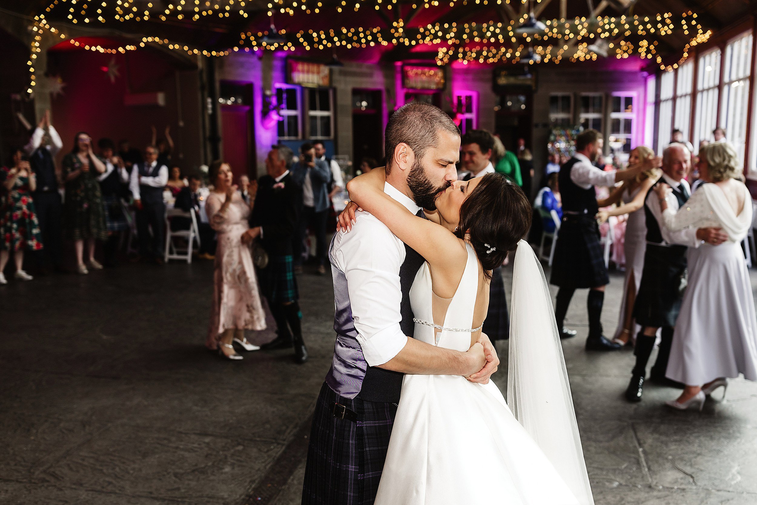 the bride and groom kiss during their first dance in a room with a stone floor and fairy lights wrapped around beams in errol park wedding venue in perthshire in scotland