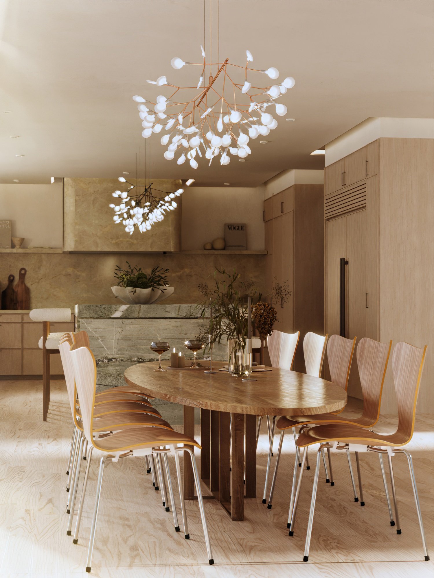 Modern dining room with a wooden oval table, light wood chairs, and decorative candles and flowers. Ceiling lights with white globes and wooden branches provide ambient lighting. The room has light wood flooring and neutral walls.