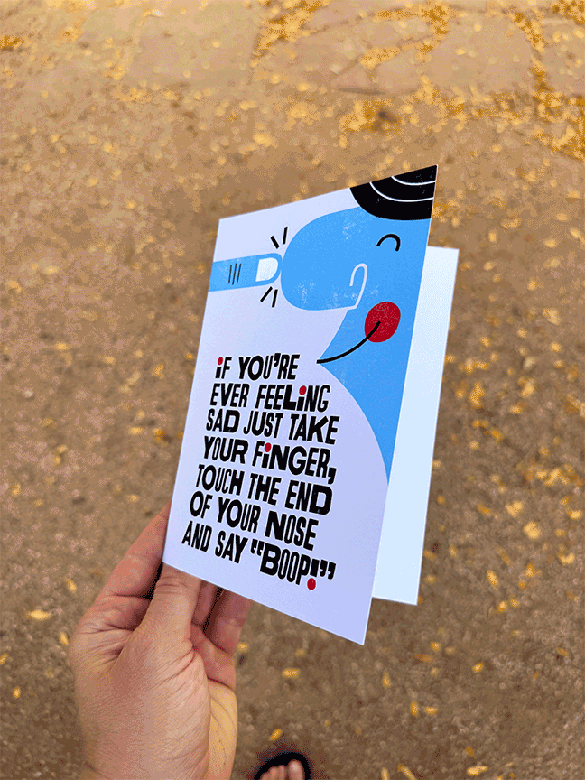 A hand holding a humorous card with an illustration of a face using a finger to touch the nose and a red dot to indicate a nose, with the text instructing to take your finger and say 'boop' if feeling sad.