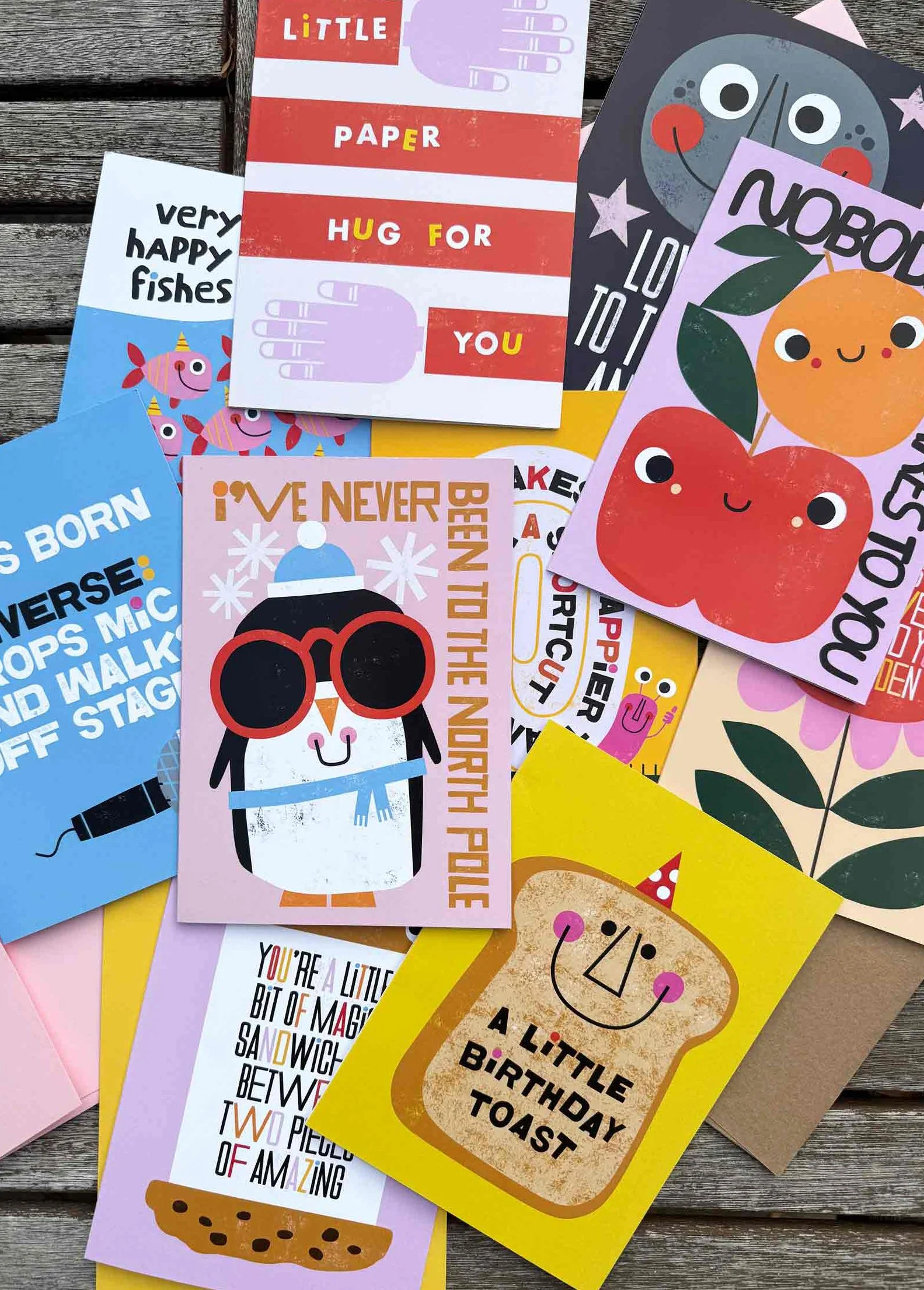 A collection of colorful birthday and greeting cards with playful designs and messages, featuring illustrations of food, animals, and cheerful characters, arranged on a wooden surface.