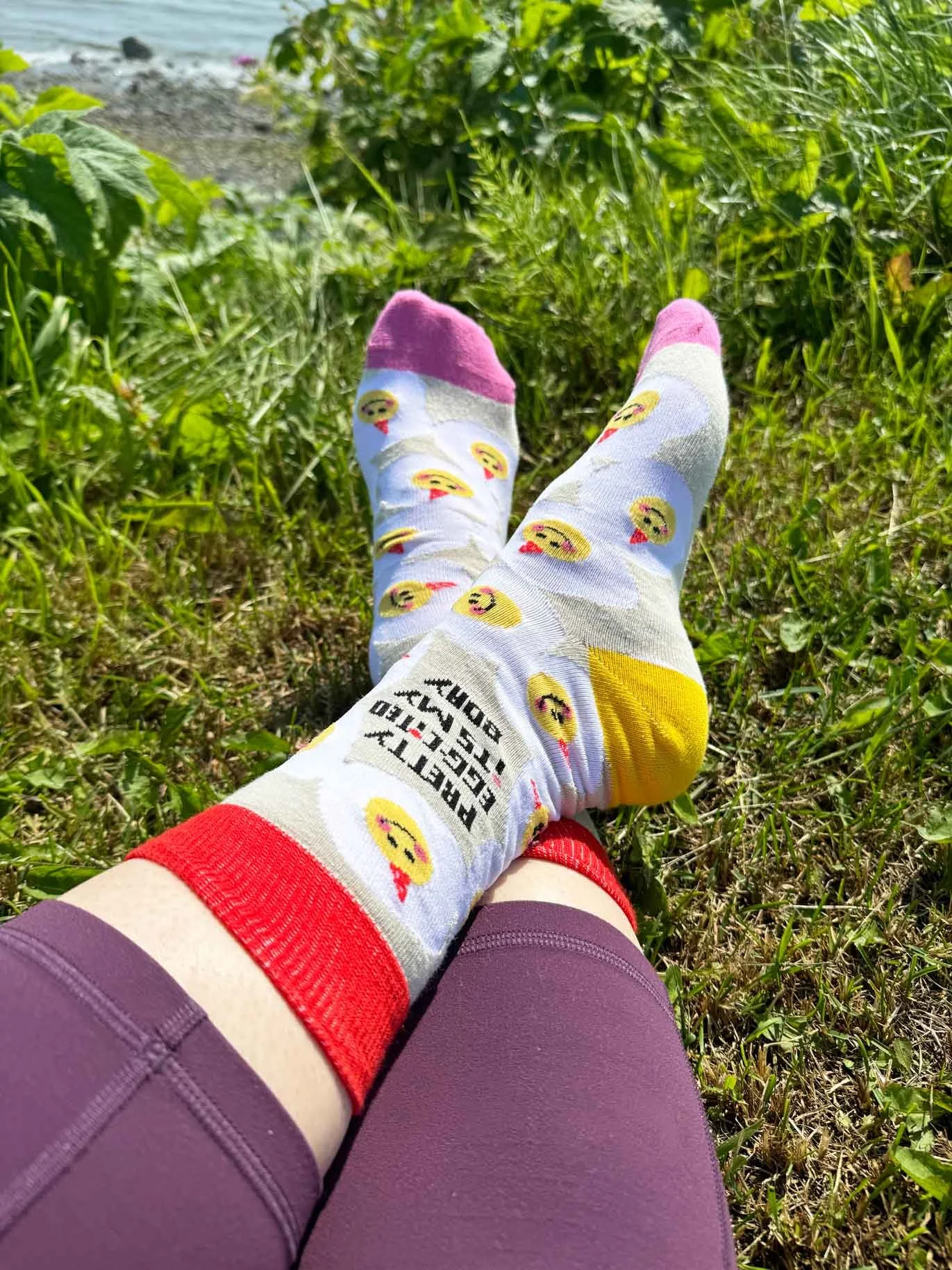 Person lying on grass near a body of water, wearing purple shorts and colorful socks with emoji faces and the phrase 'Pretty Egg-cited It's My BDay' written on one sock.