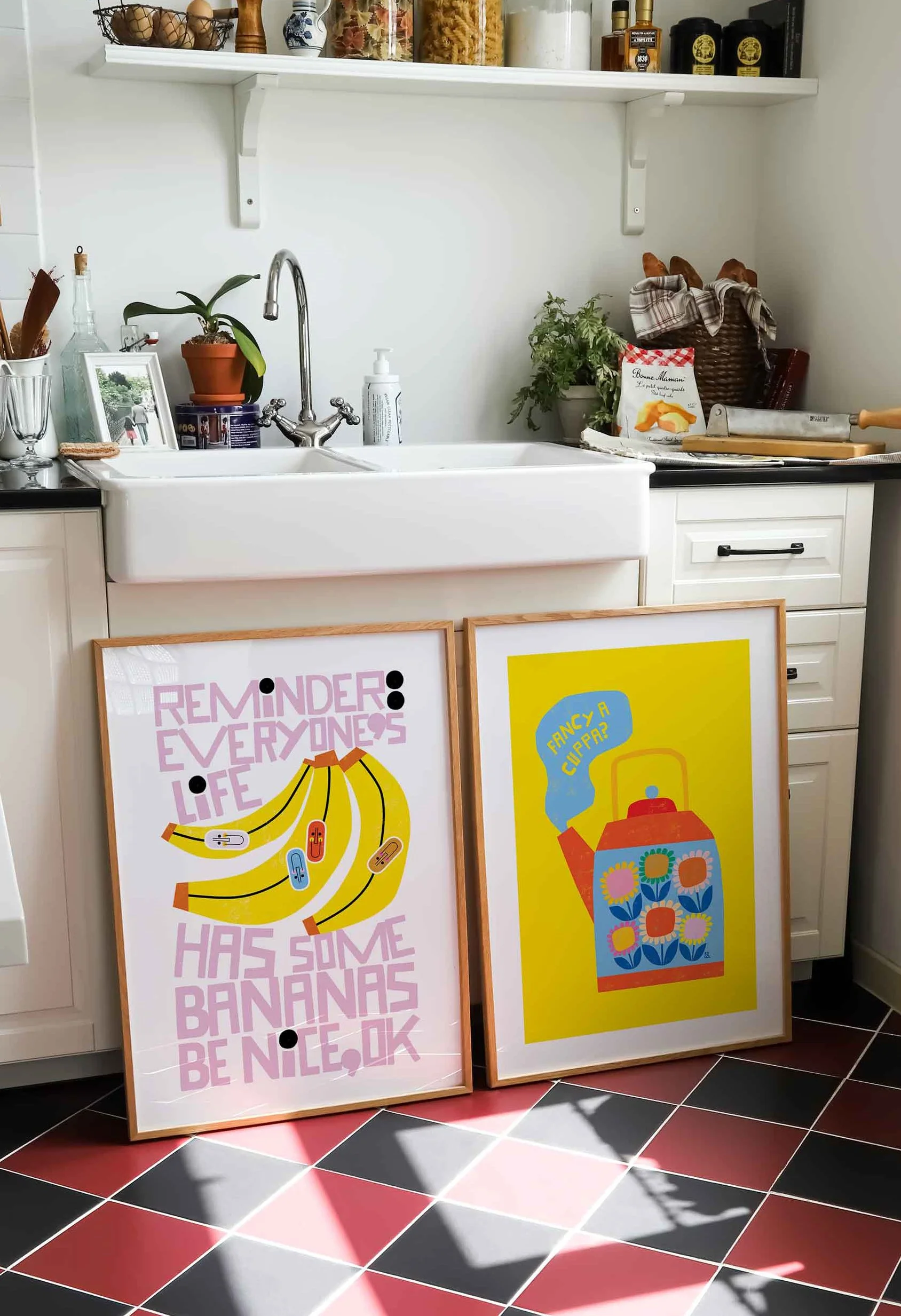 Two framed prints lean against a white kitchen sink and cabinet. The left print features pink text and a cartoon banana with the message, "Reminder: Everyone's life has some bananas be nice, ok." The right print displays a colorful teapot with a spee