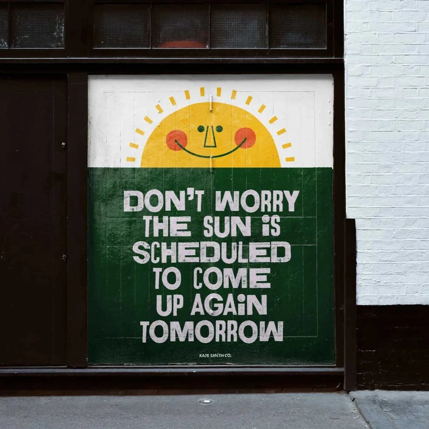 A sign on a wooden board with a cheerful sun illustration and the text, 'Don't worry, the sun is scheduled to come up again tomorrow,' attributed to Kate Smith Co.
