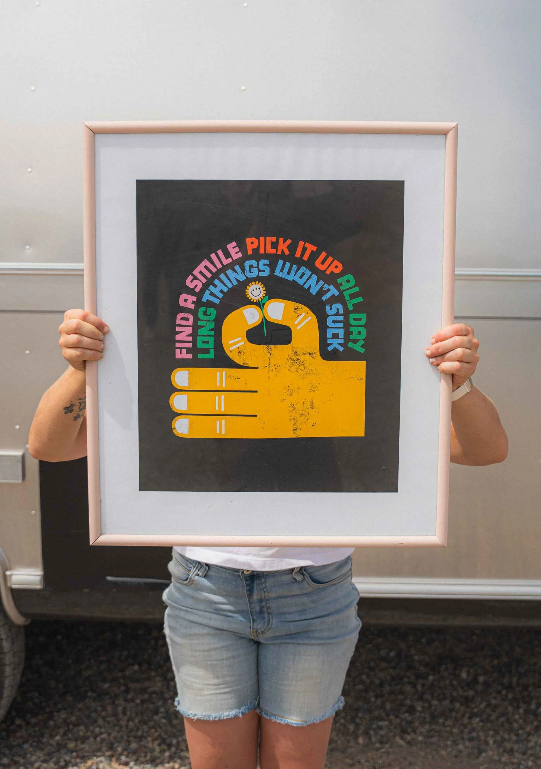 Person holding a framed poster with a yellow cartoon hand, a black face with a straw, and colorful text that reads 'Find a smile pick it up all day long things won't suck.'