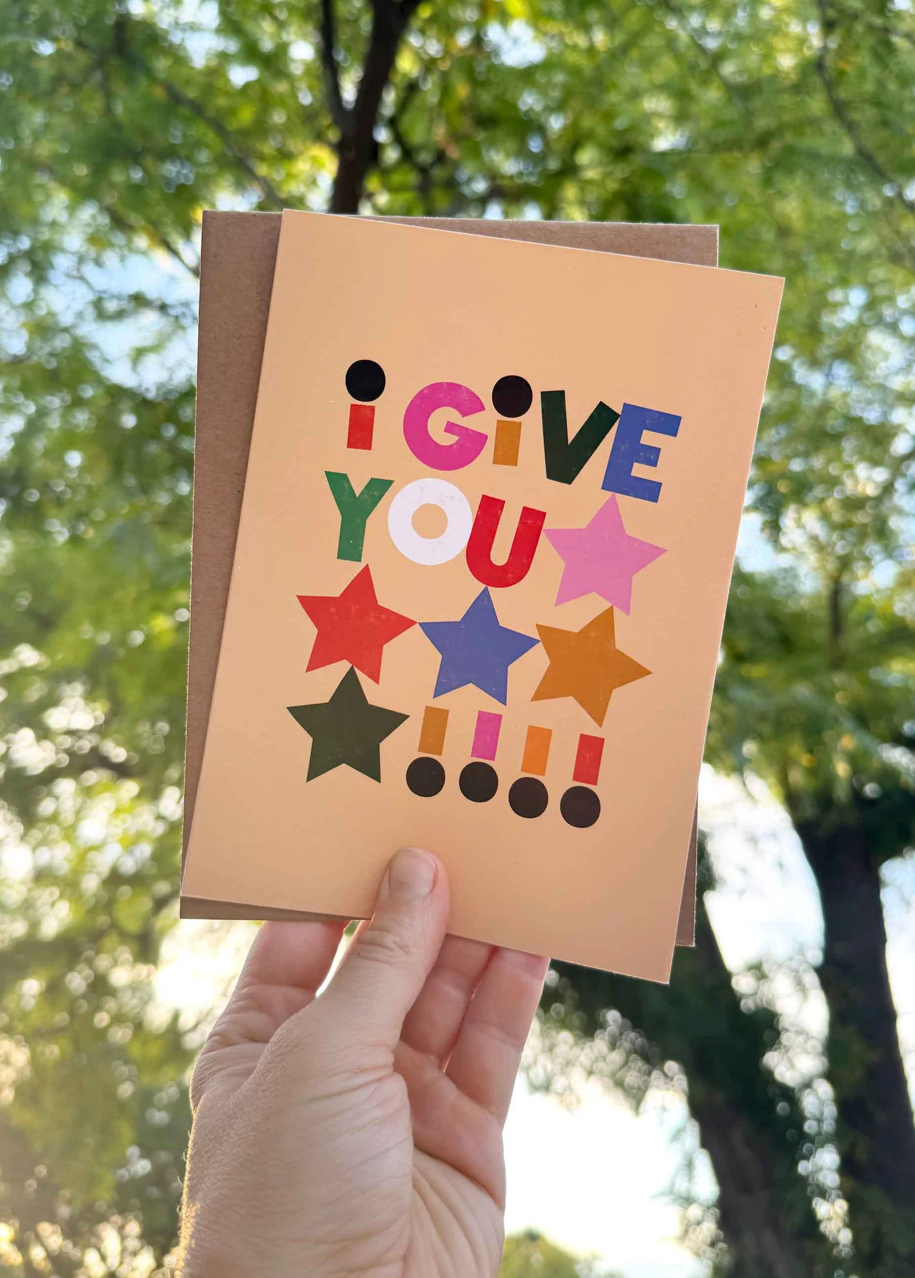 Hand holding a colorful greeting card with the text 'I GIVE YOU !!!' and various star shapes, outdoors with trees and green leaves background.