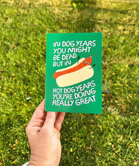 A hand holding a humorous greeting card outdoors on a grassy area. The card features a cartoon hot dog with a smiling face, sunglasses, and a party hat, along with the text 'In dog years you might be dead but in hot dog years you're doing really grea