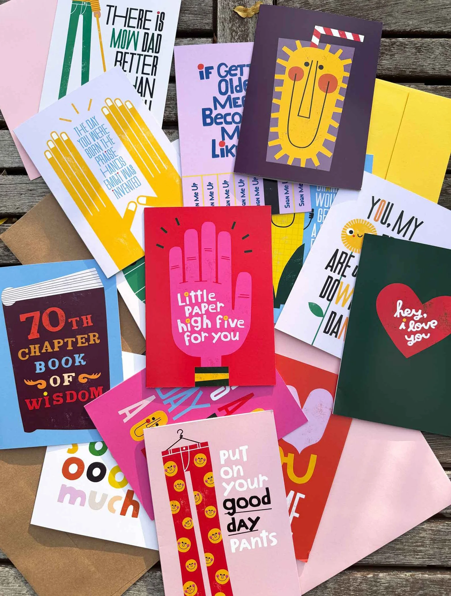 A collection of colorful greeting cards and note cards with uplifting messages, some featuring smiley faces, hearts, and playful illustrations, spread out on a wooden surface.