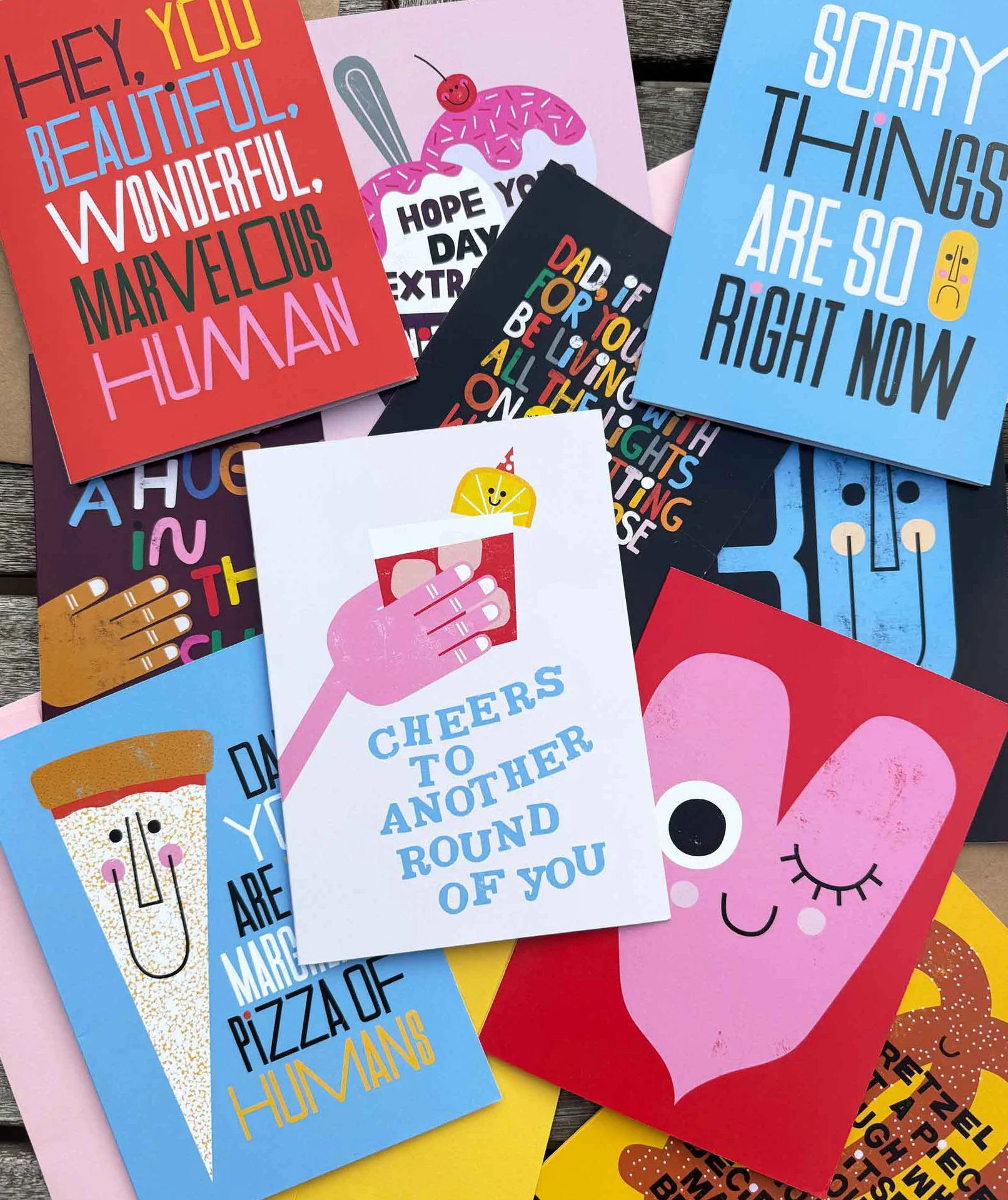 A collection of colorful greeting cards with playful messages and illustrations, including a pink heart with a face, a slice of pizza with a face, and various cheerful phrases.