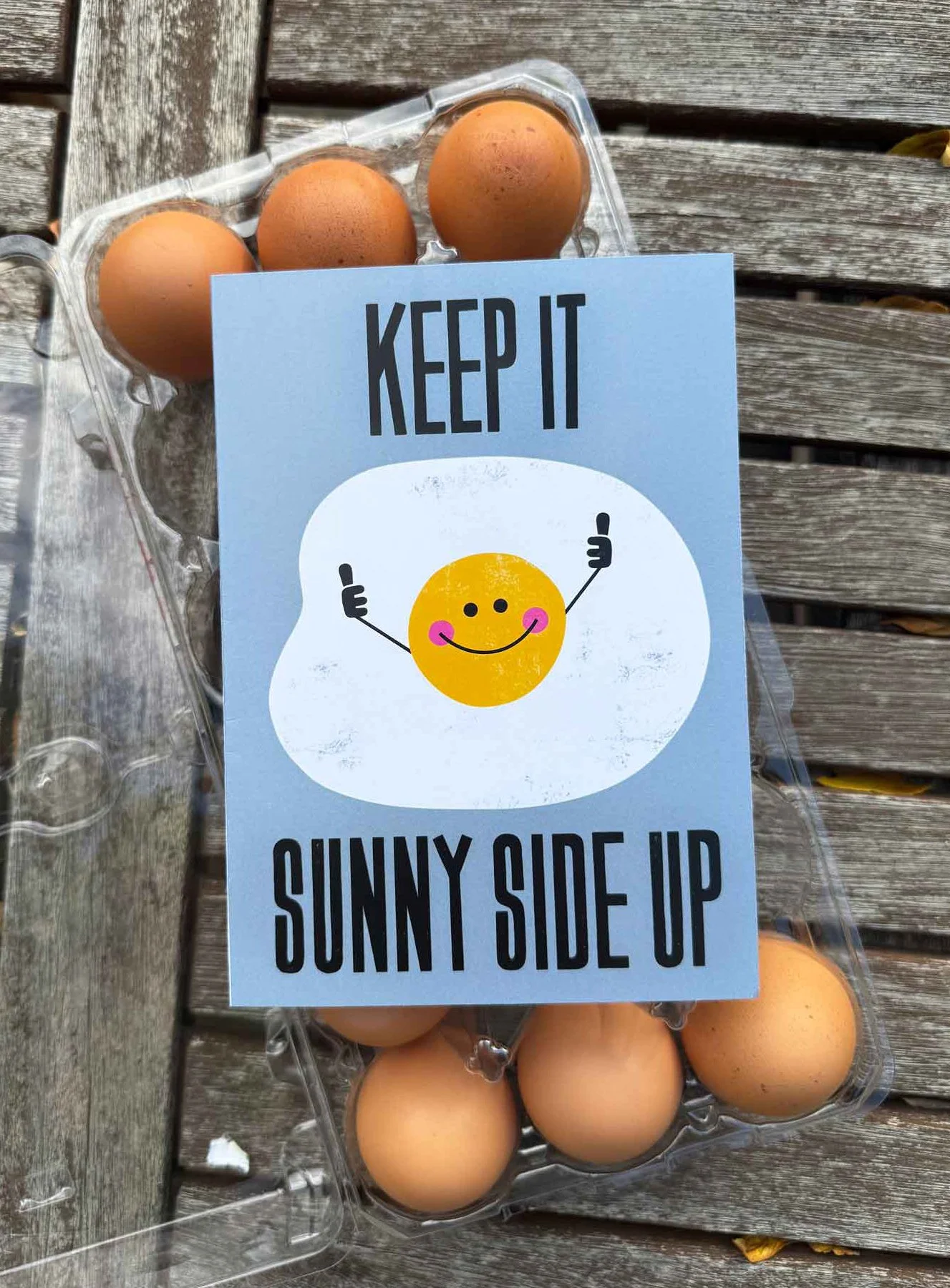 A carton of brown eggs with a greeting card on top that says "Keep it sunny side up" with a smiling sun illustration.
