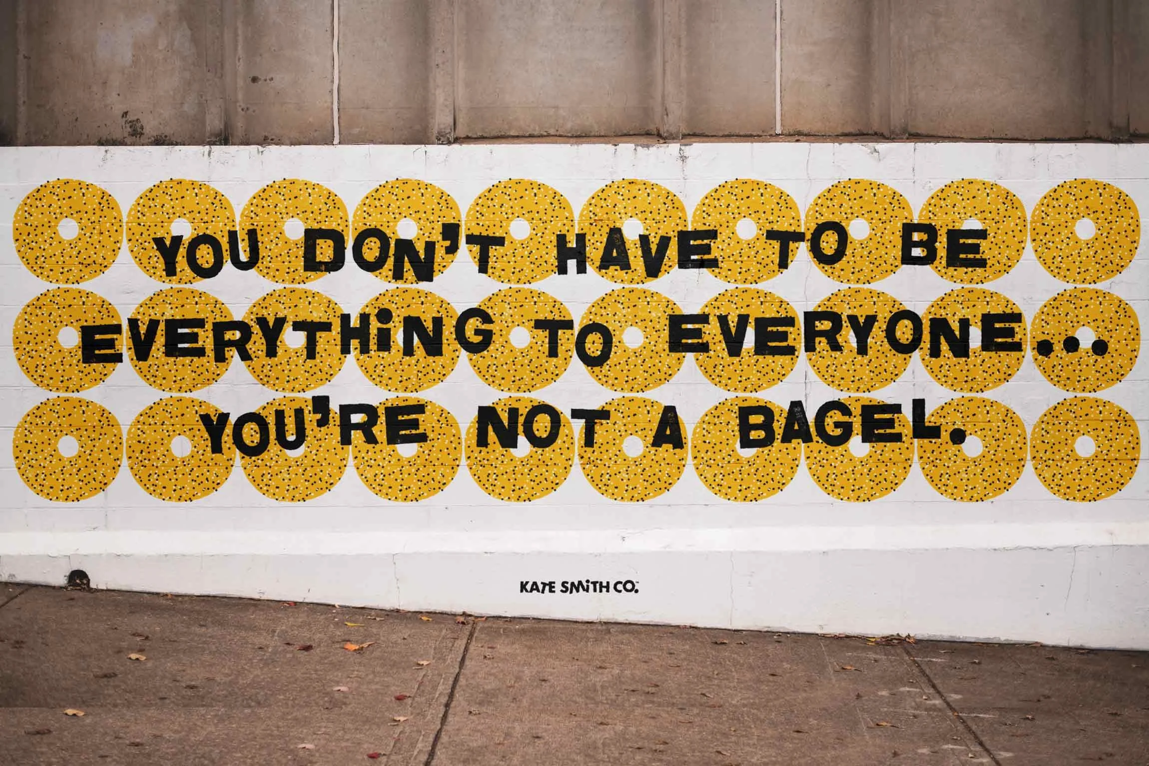 Wall mural with yellow bagel illustrations and black text saying, "You don't have to be everything to everyone... You're not a bagel" by Kate Smith Co.