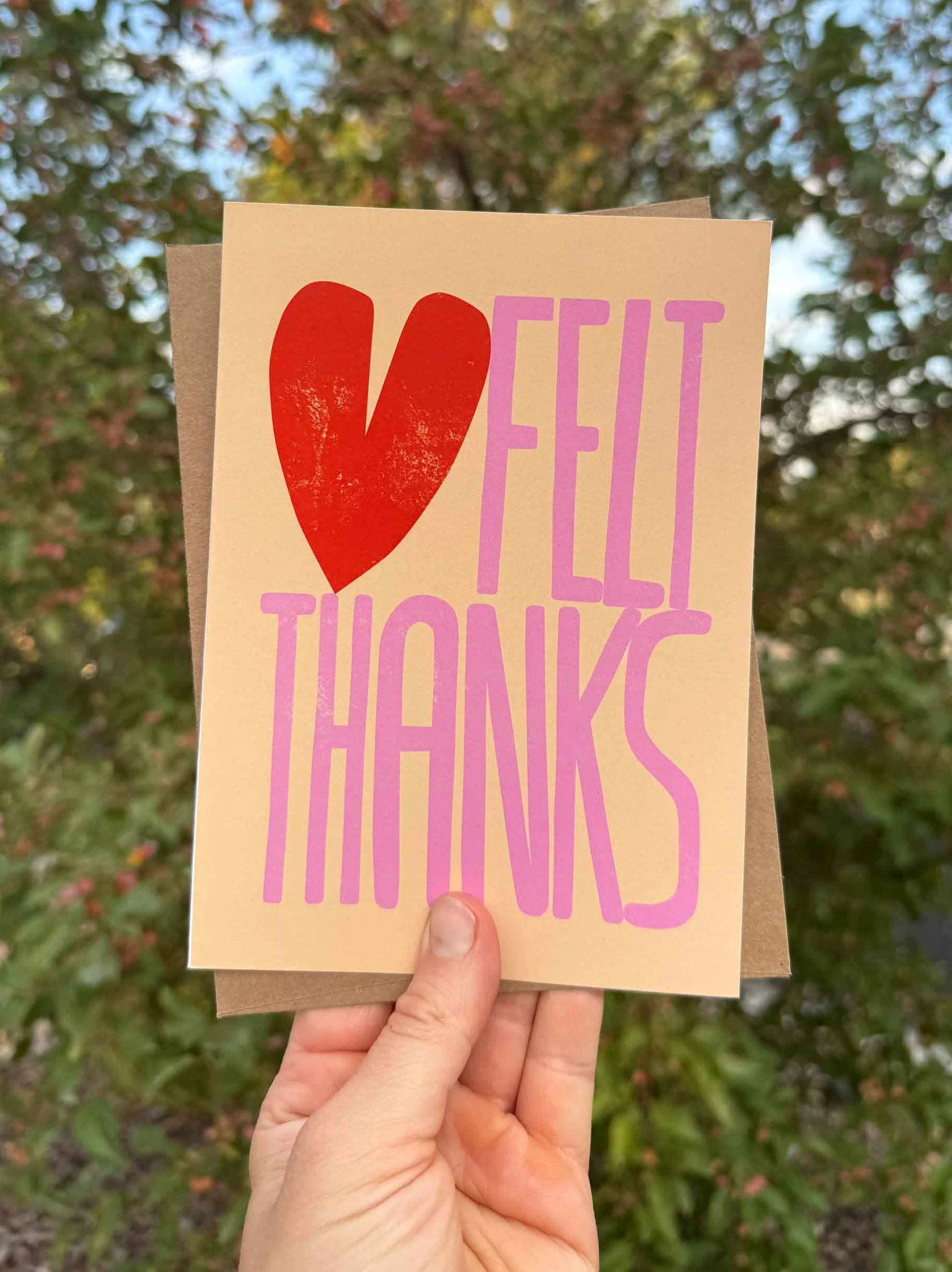 A hand holding a card with the words 'FELL THANKS' and a red heart on a background of trees with fall foliage.