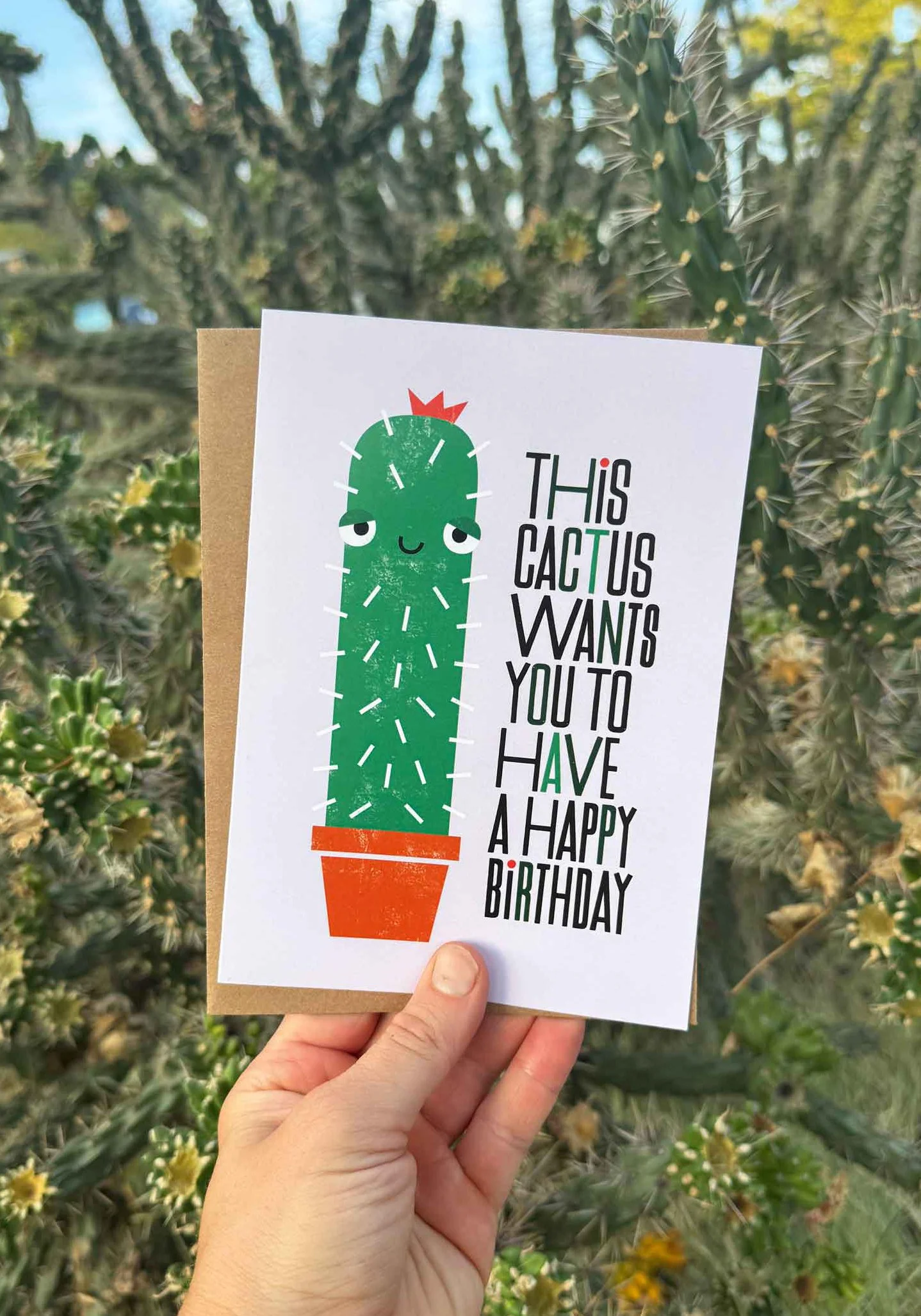 A birthday card with a cartoon cactus drawing and the message: "This cactus wants you to have a happy birthday," held in a person's hand against a background of cactus plants.
