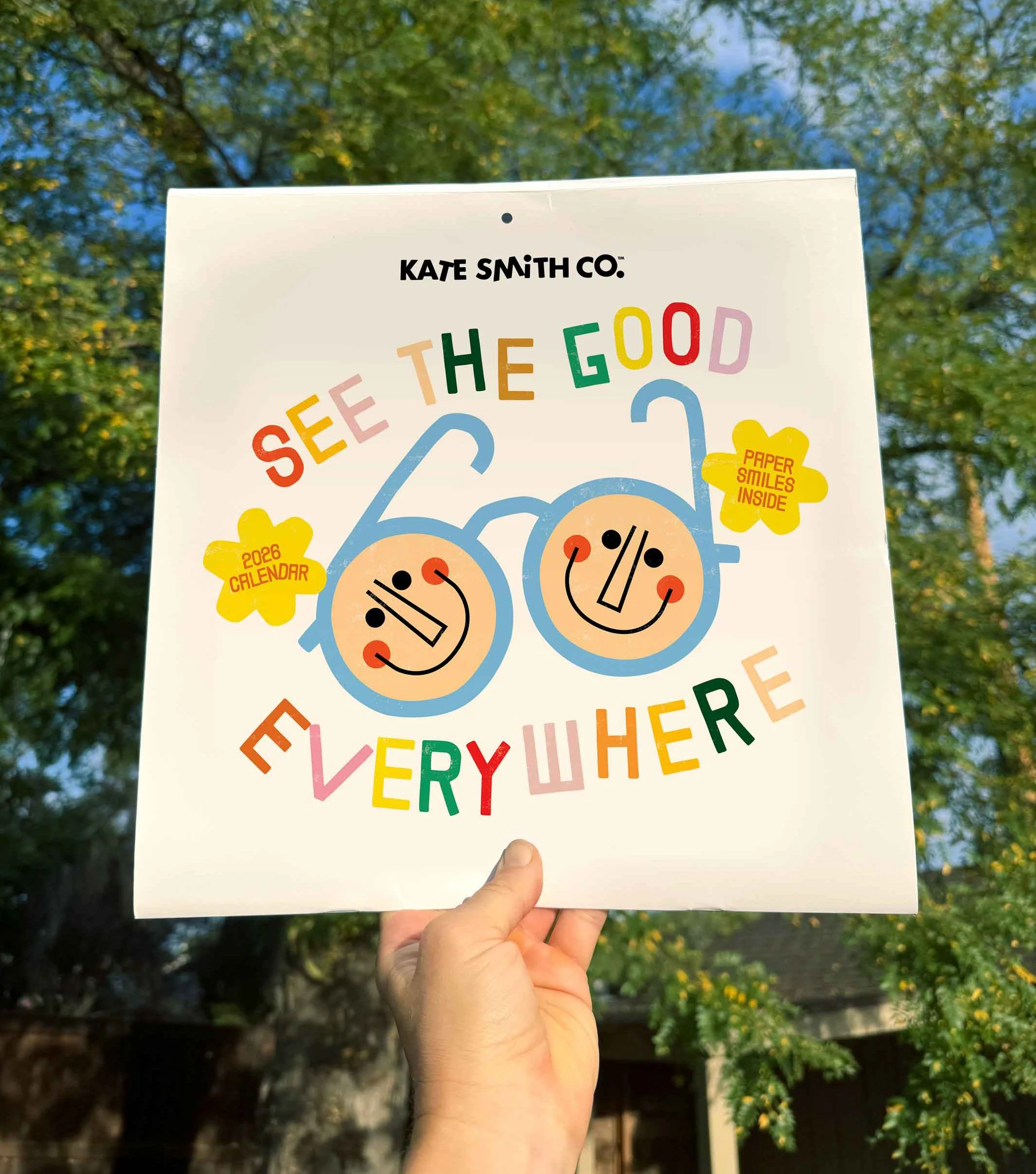Calendar cover titled 'See the Good Everywhere' by Kate Smith Co., featuring a colorful illustration of glasses with smiley faces, yellow speech bubbles with '2026 Calendar' and 'Paper Smiles Inside,' and multicolored text.