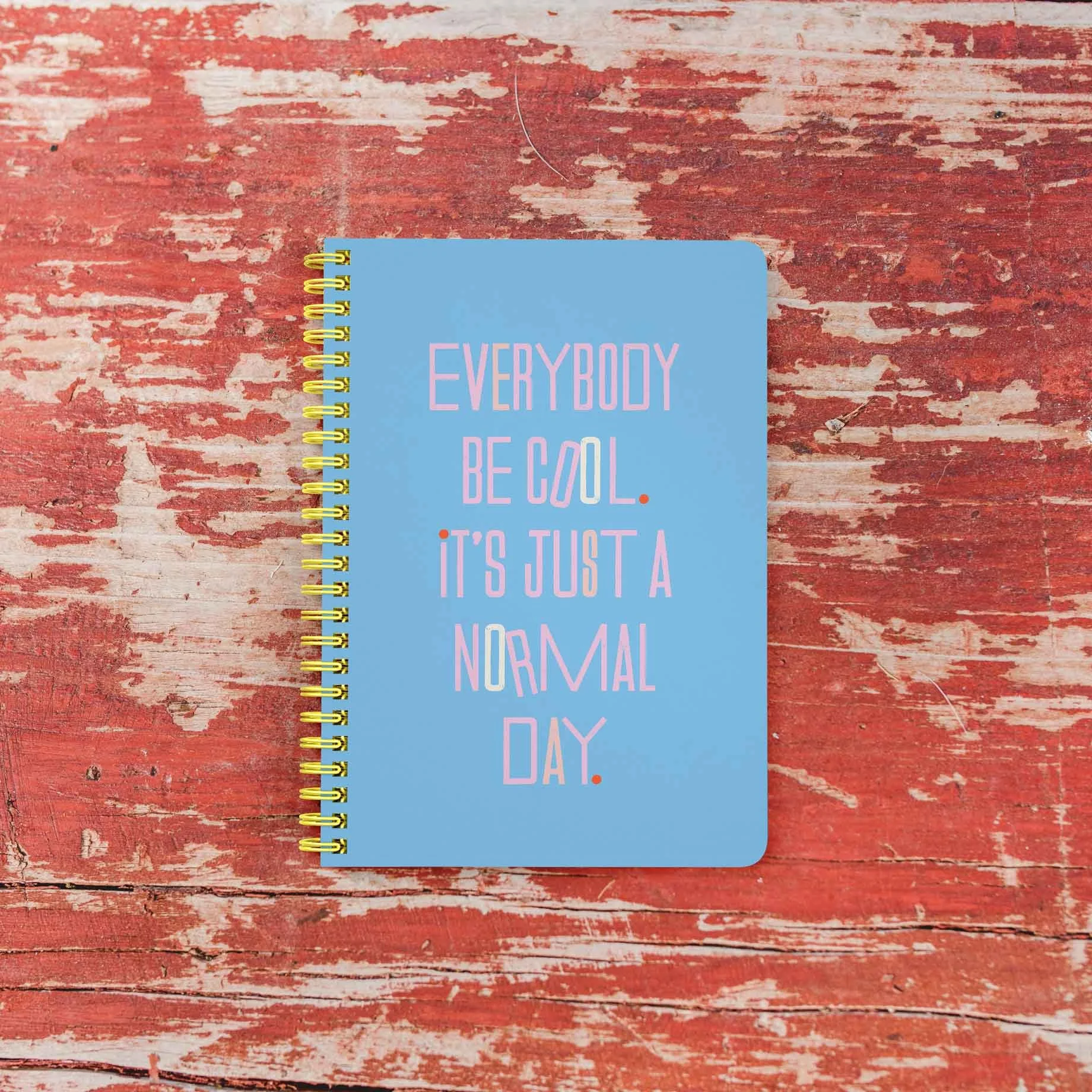 A blue spiral notebook with the phrase "Everybody be cool. It's just a normal day" written in pink, white, and multicolored text, placed on a weathered red wooden surface.