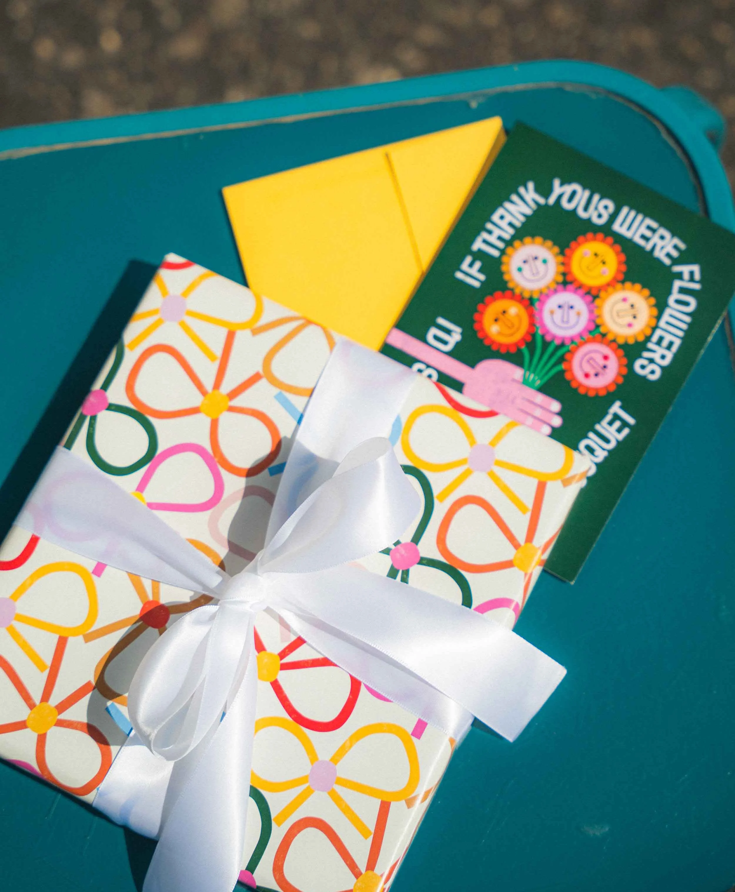A colorful gift wrapped in flower-patterned paper with a white ribbon bow, resting on a teal surface, with a greeting card and a small yellow card on top.