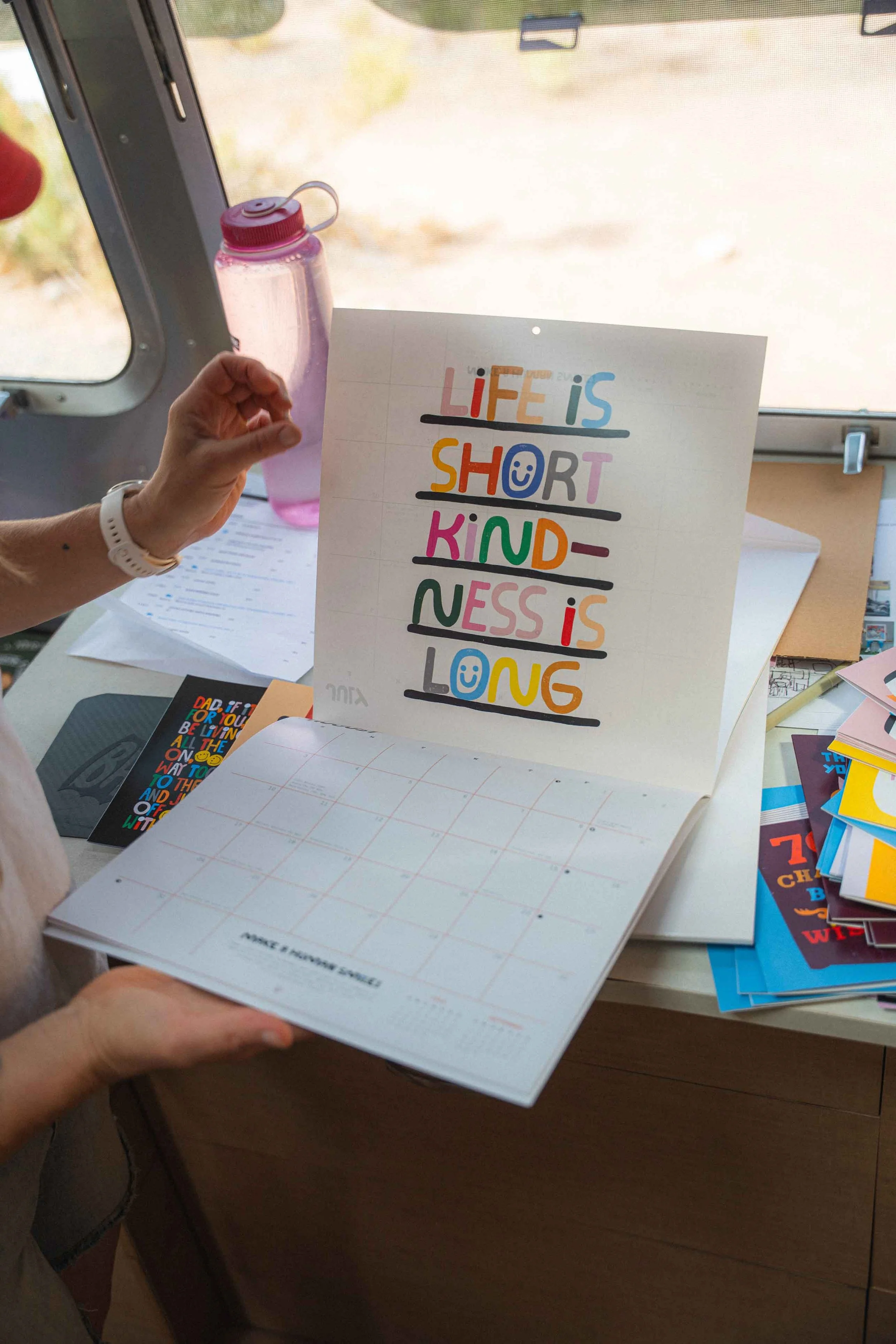 Colorful sign that reads 'LIFE IS SHORT, KINDNESS IS LONG' with smiley faces and underlines, held by a person near a window with a pink water bottle on the windowsill, and a table with various papers and cards nearby.