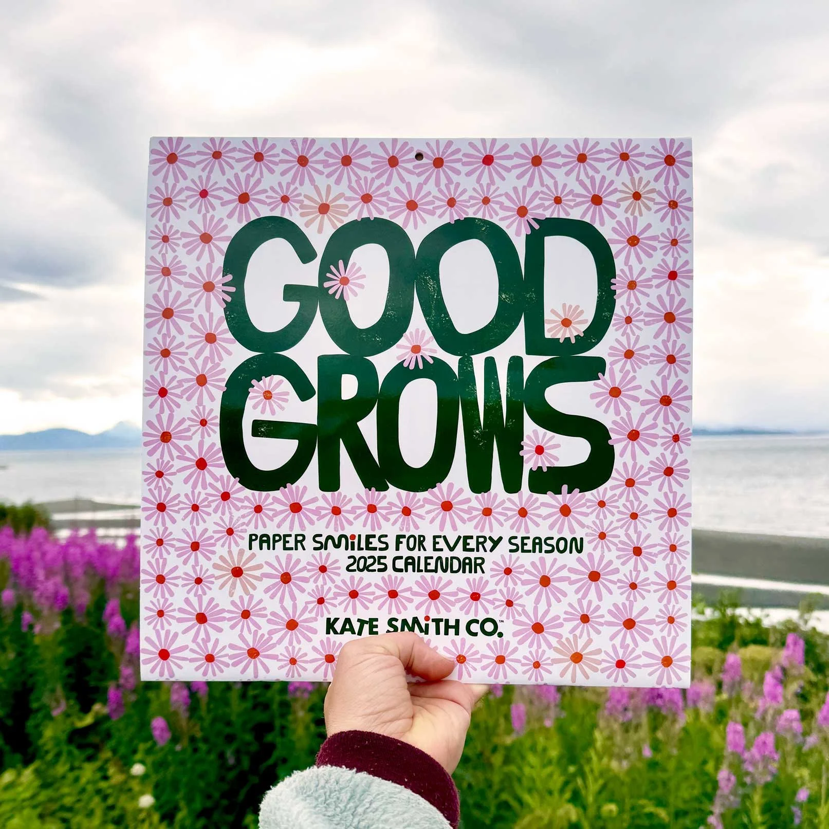 Hand holding a 2025 calendar titled 'Good Grows' with a pink flower pattern, against a background of purple flowers, grass, water, and mountains.