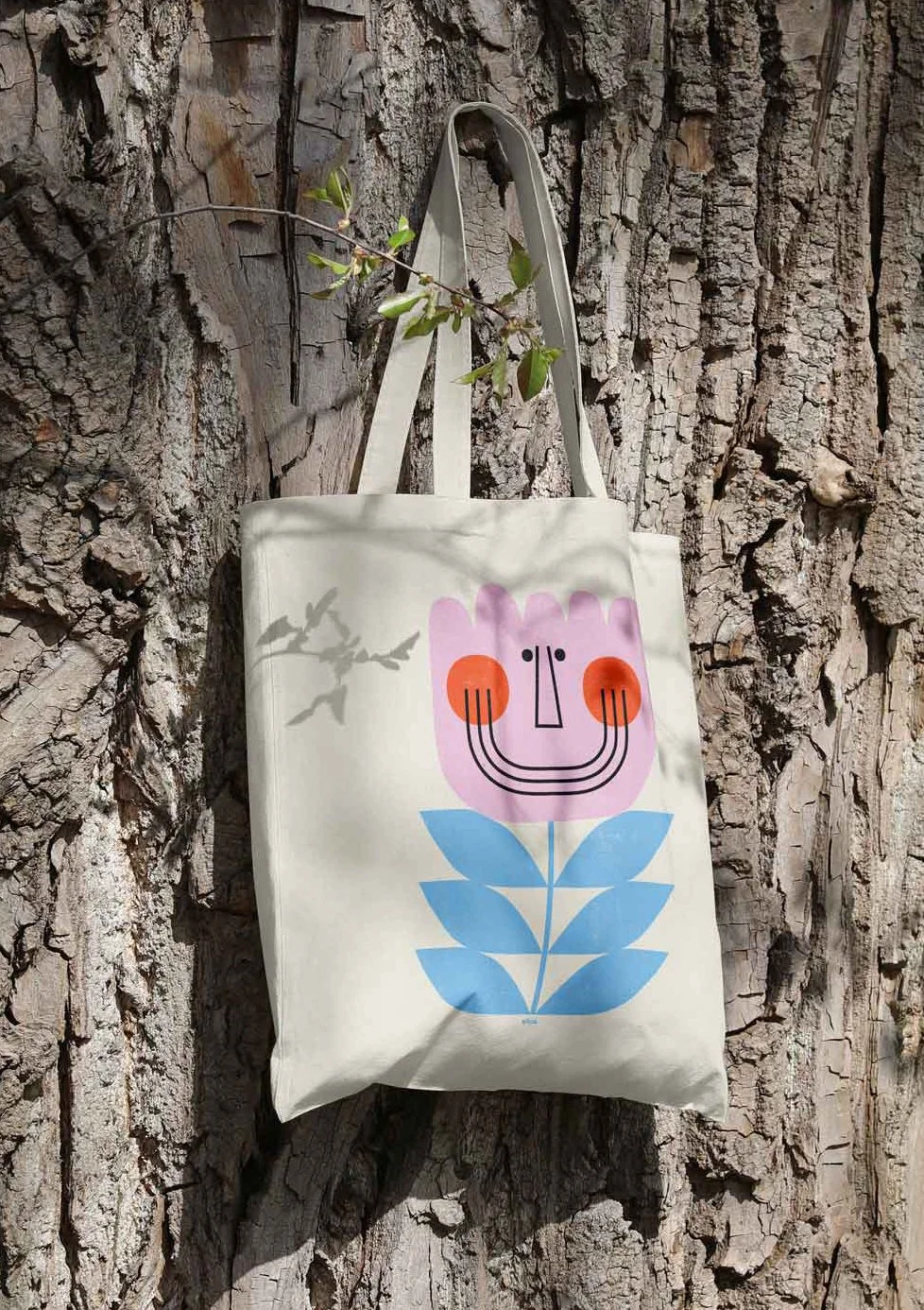 A white tote bag with a colorful, abstract face and leaf design, hanging on a rough tree bark background with a small branch and leaves casting a shadow.