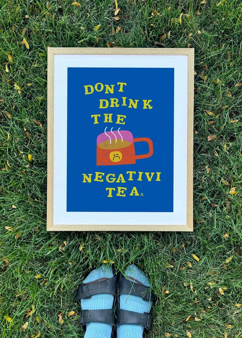 A colorful sign with a humorous message about avoiding negative tea, featuring a cartoon tea mug with a sad face, on grass with a person's feet in sandals and socks visible at the bottom.