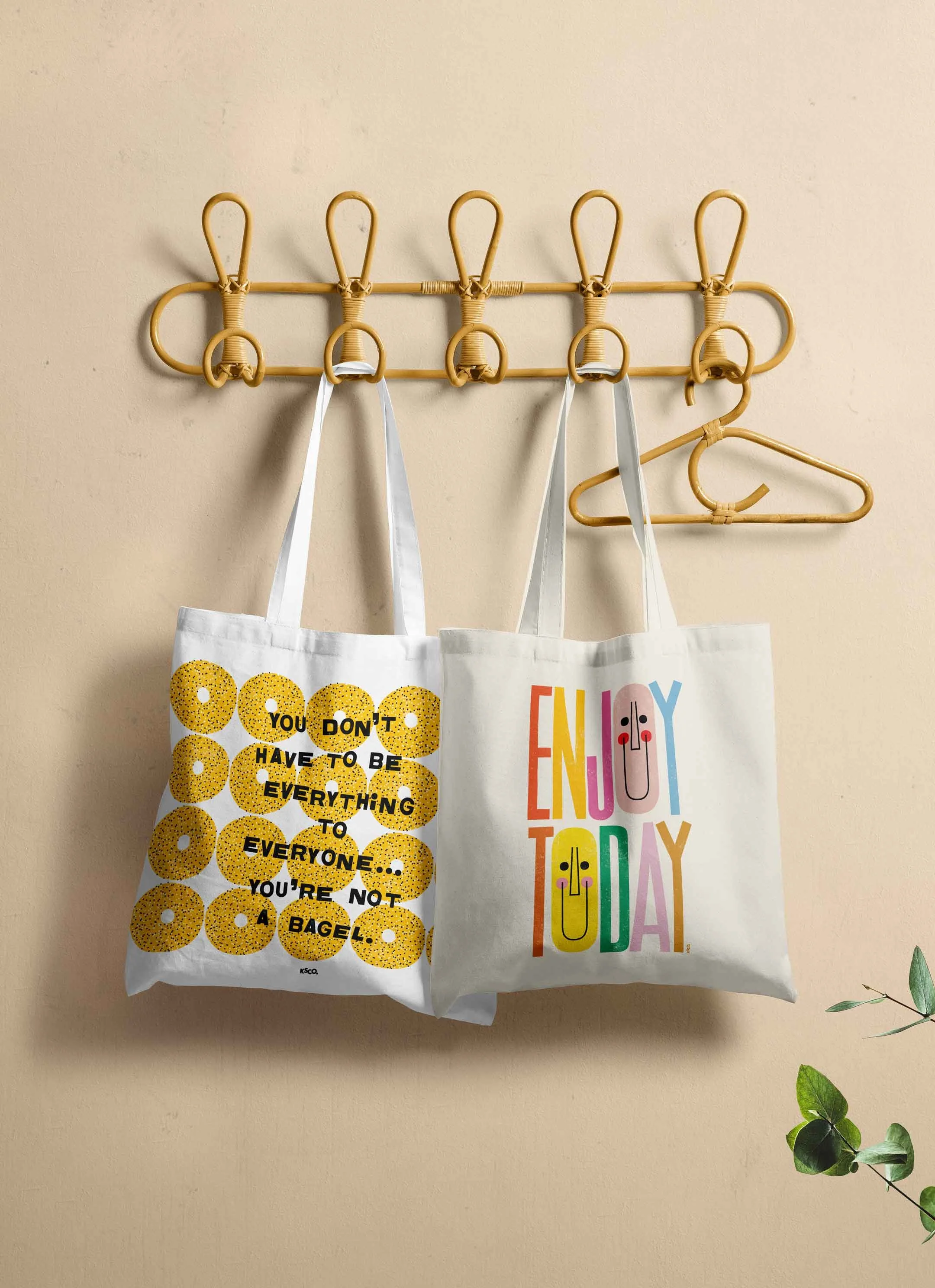 Two reusable tote bags hanging on a beige wall with a wooden rack. The bag on the left has a yellow donut pattern with black text that reads, 'You don't have to be everything to everyone... you're not a bagel.' The bag on the right has colorful text