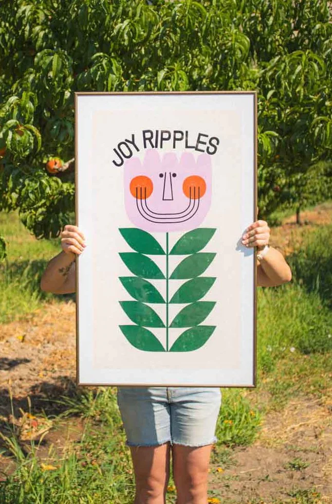 Person holding a framed artwork with a stylized smiling flower face and green leaves, the words 'Joy Ripples' written above, set outdoors with trees and greenery in the background.