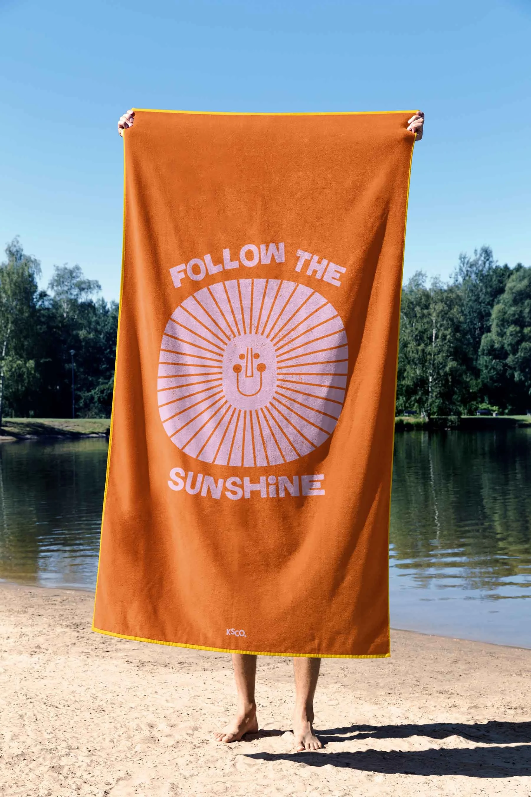 Person holding an orange towel with the logo and text 'Follow the Sunshine' by a lakeside during daytime.