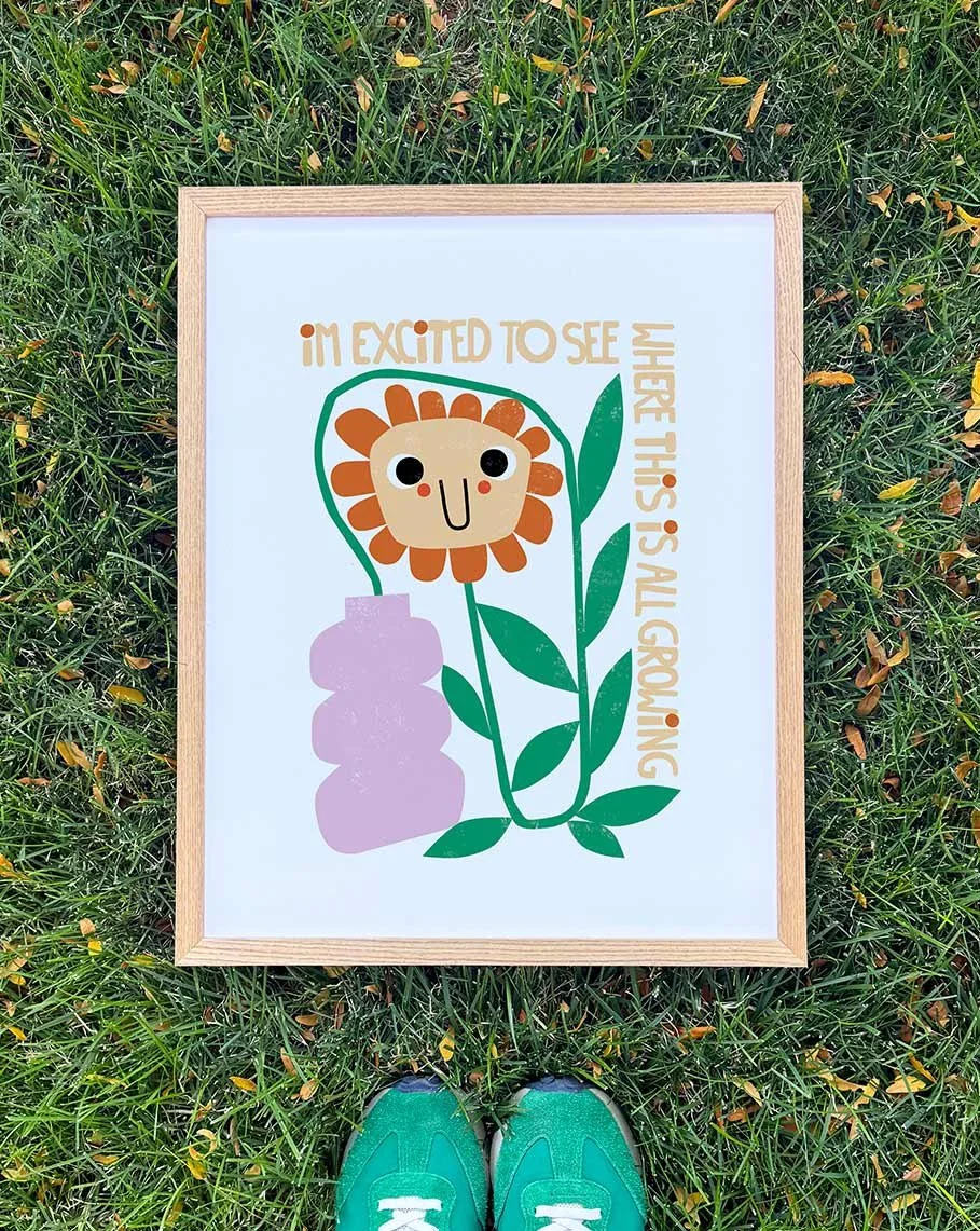 A framed illustration of a smiling flower with a face, surrounded by green leaves and a pink rock, with the text "I'm excited to see where this is all growing".