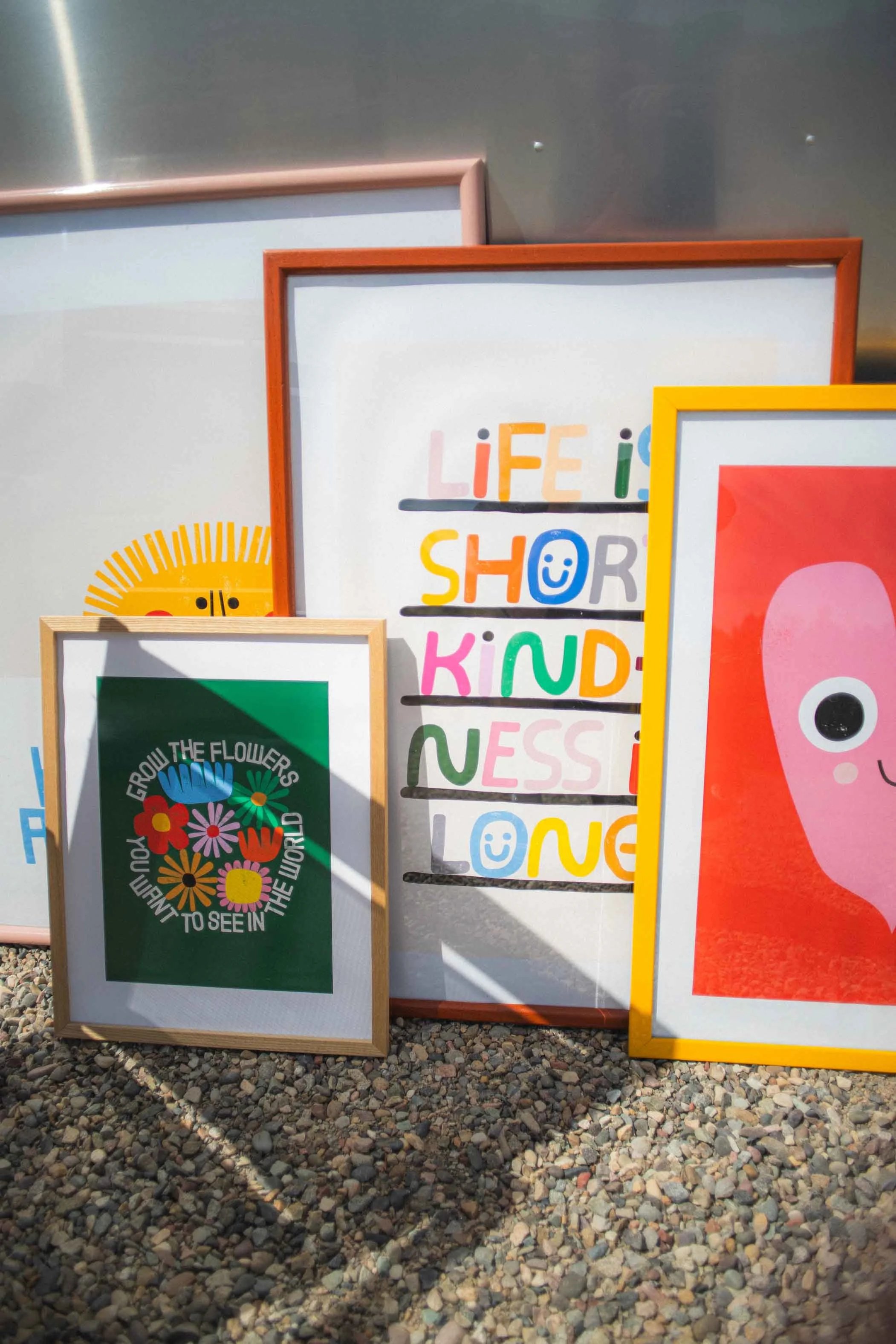 A collection of colorful framed prints with positive and inspiring messages, including one that says 'LIFE IS SHORT KINDNESS IS LONG,' arranged outdoors against a metallic wall on a gravel ground.