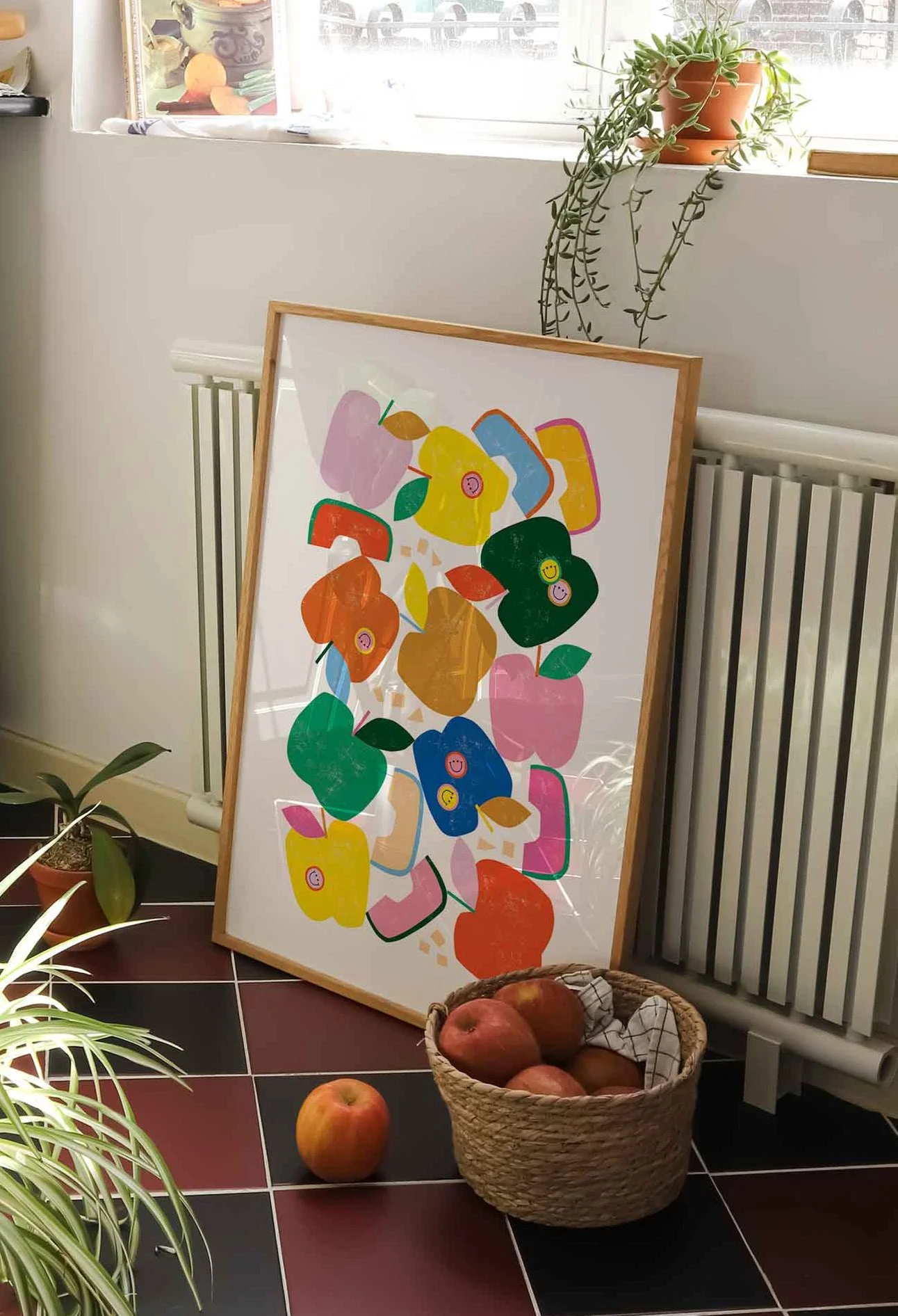 A colorful abstract art print leaning against a wall near a white radiator, with a basket of apples on the floor and a potted plant on the windowsill.