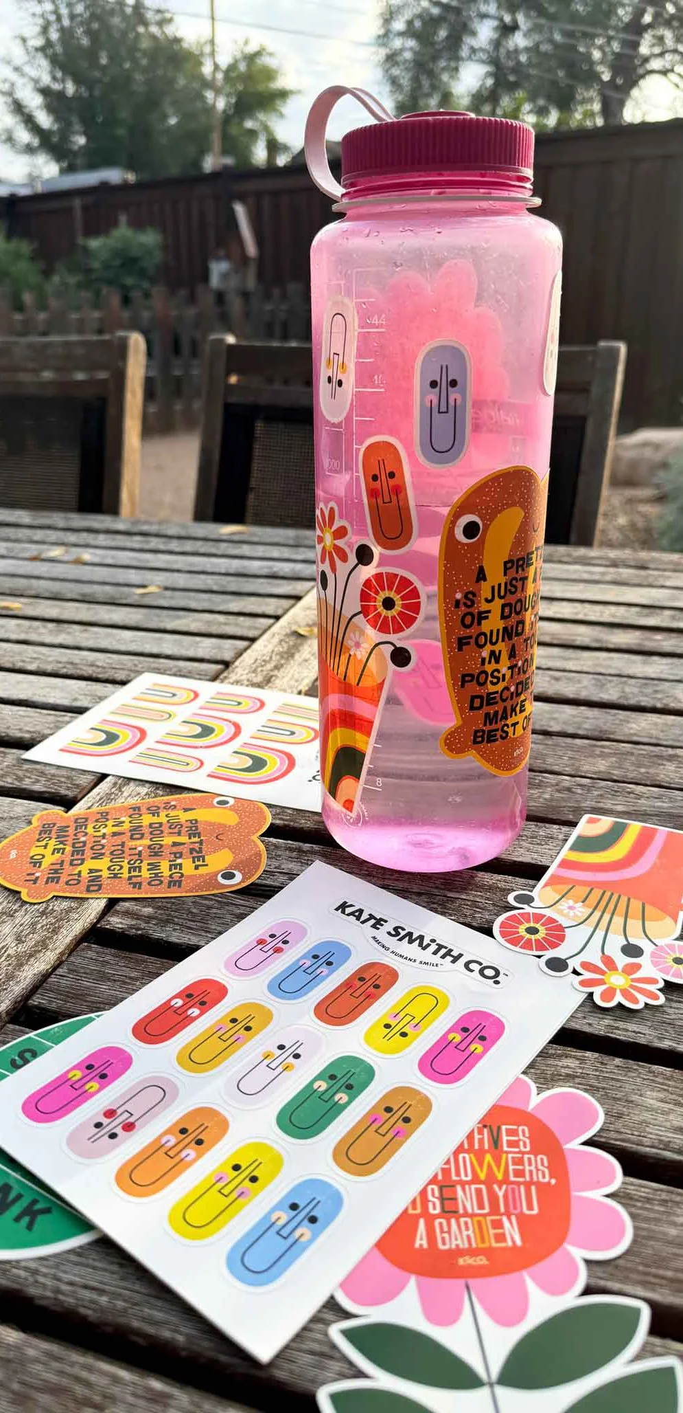 A pink water bottle with colorful cartoon faces and a sticker with an orange creature and a quote. The bottle is on a wooden table with colorful stickers, a sheet of colorful face stickers, and floral decorative items scattered around. Outdoor settin