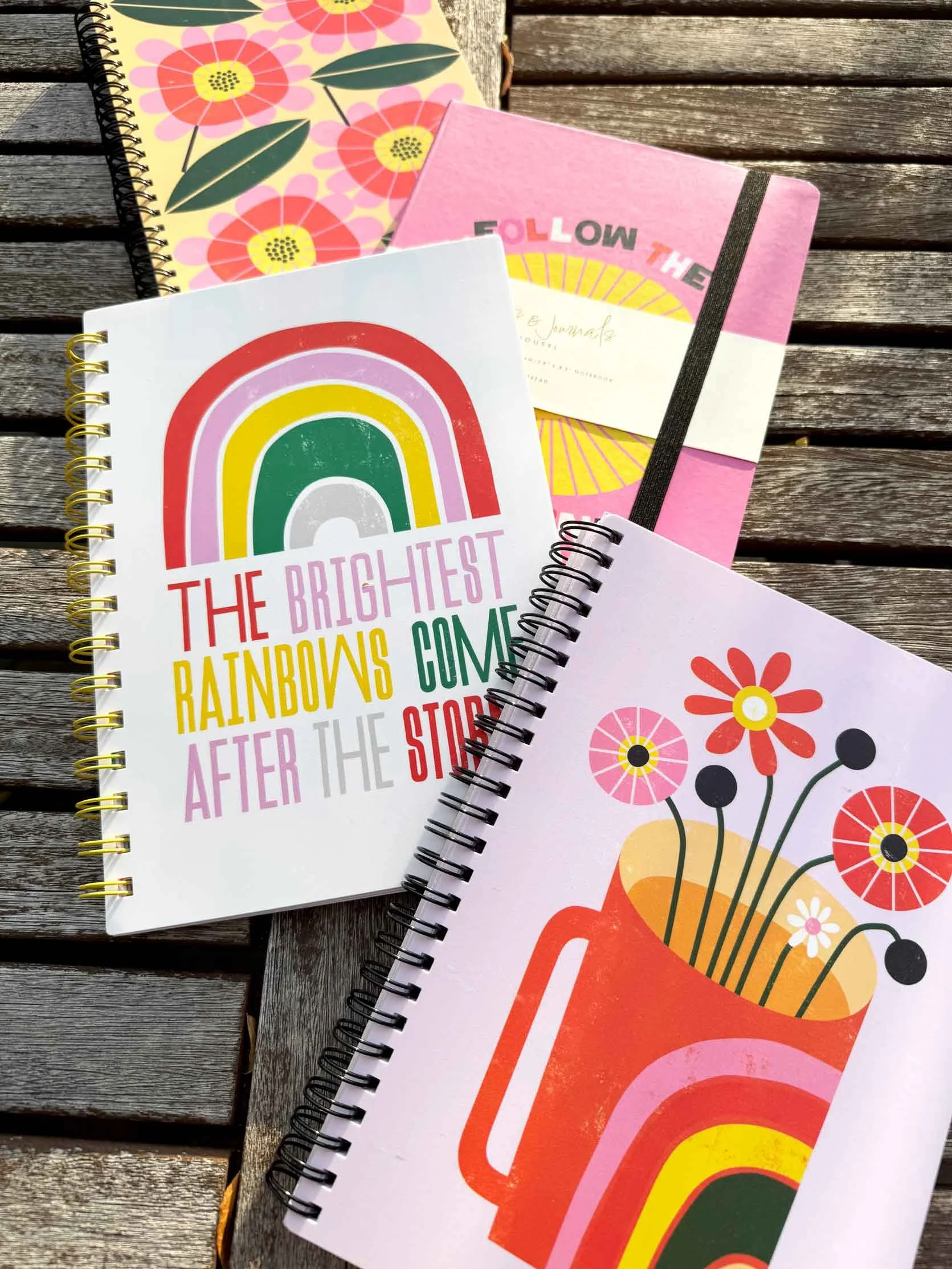 Four colorful spiral-bound notebooks with floral and rainbow designs on a wooden surface.