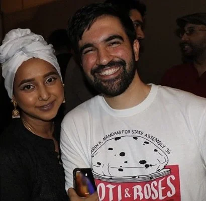 A woman with a white headwrap and a man with a beard smiling for a selfie at a social event.