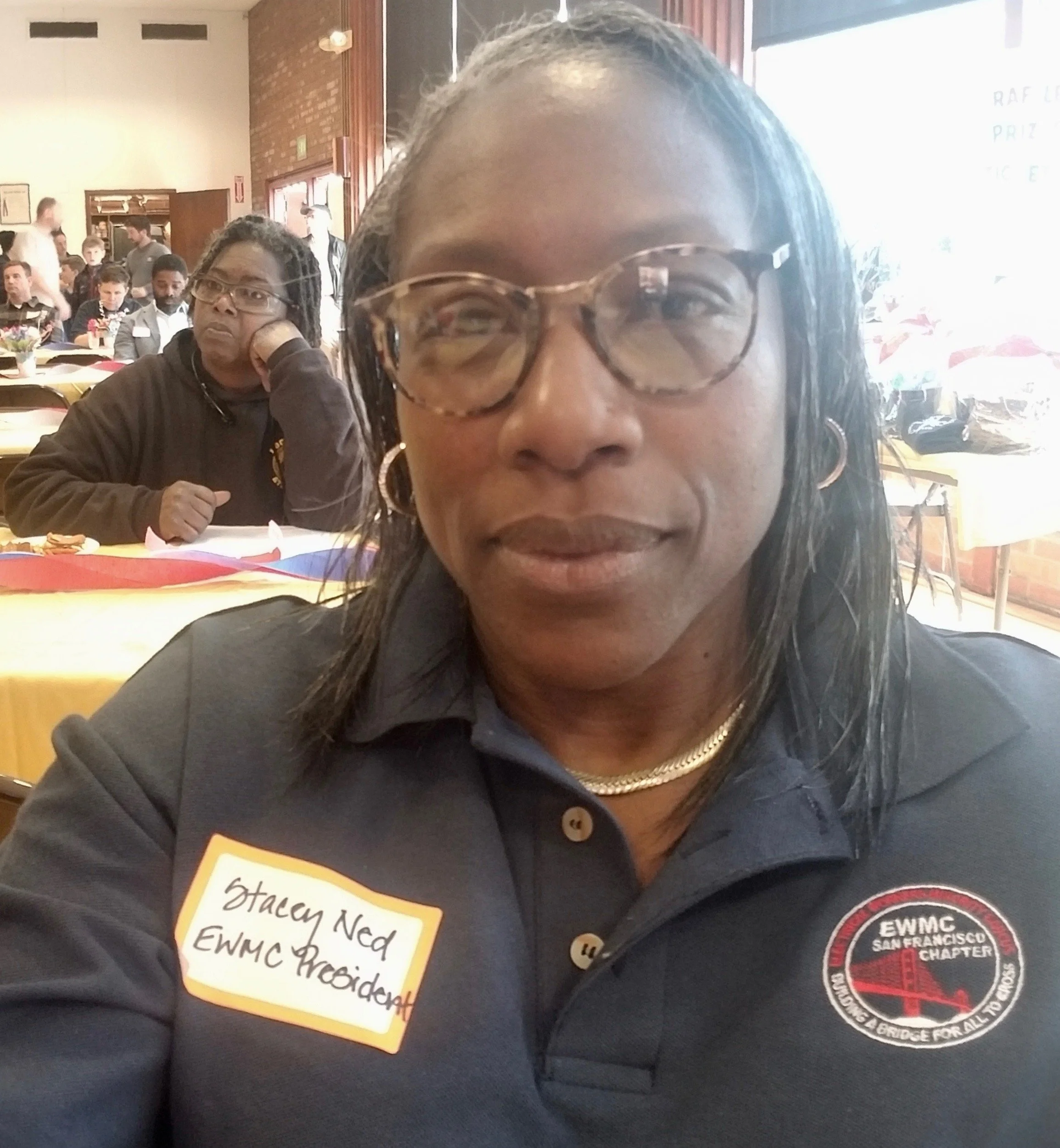 A woman wearing glasses and a dark uniform with a badge and a patch that reads "EWMC SAN FRANCISCO CHAPTER" is in the foreground. Behind her, a man with glasses and dreadlocks, wearing a similar uniform, is sitting at a table with colorful tablecloth