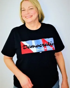 Smiling woman wearing a black T-shirt with a Democratic logo and slogan in front of a plain background.