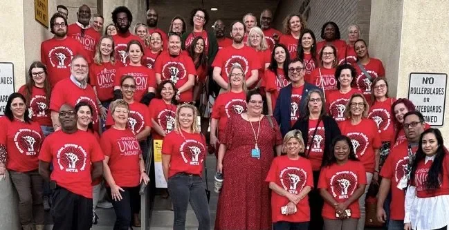 Group of diverse people from a teachers union standing together, wearing red shirts with a logo, posing for a photo outside a building.
