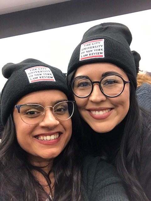 Two women wearing black beanies with the logo for The City University of New York Law Review, smiling and taking a selfie indoors.
