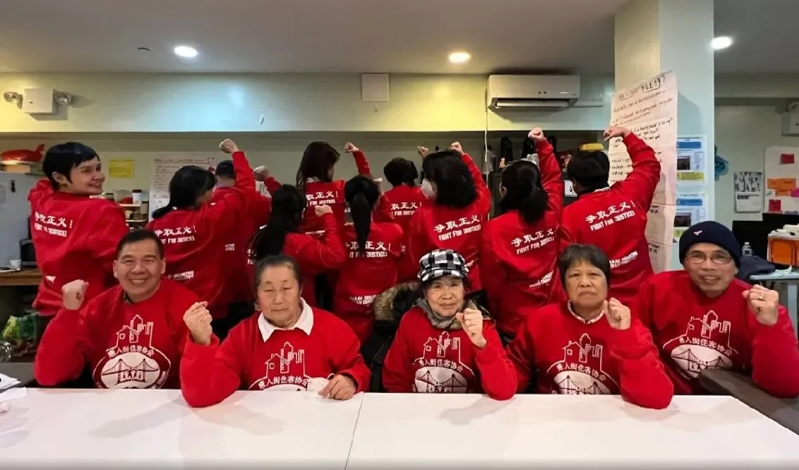 Group of people, mostly elderly, sitting at a table and wearing red shirts with white graphics, posing with clenched fists. Others in red shirts are standing behind them, some with raised fists, in a room with posters and an air conditioner.