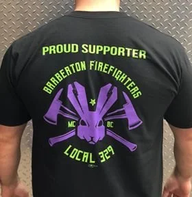 Back of a black T-shirt with green and purple text and graphics supporting Berberton firefighters. The text reads 'PROUD SUPPORTER', 'BERBERTON FIREFIGHTERS', 'LOCAL 329', and has a purple logo of a firefighter's helmet with two axes crossed behind i