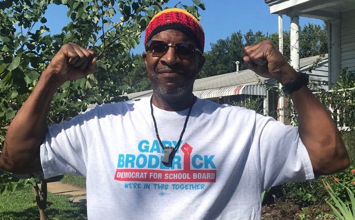 Man flexing his biceps outdoors, wearing sunglasses and a campaign T-shirt supporting Gady Broderick for school board.