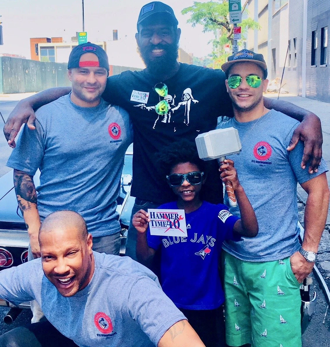 Group of five peopl posing together outdoors, smiling, with city buildings and trees in the background, two men wearing gray T-shirts with a red logo, a young boy holding a sign that says "Hammer Time", 