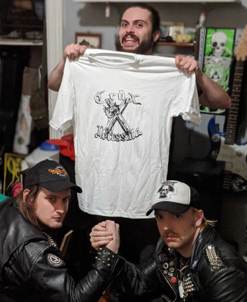 Three men in a room, with one standing behind and holding a white T-shirt with a 'Grixx Neussala' logo, and two others kneeling in front, engaging in an arm-wrestling match, wearing leather jackets and baseball caps.