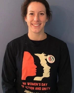 A woman smiling, wearing a black T-shirt with a Women's Day graphic, standing against a plain wall.