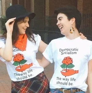 Two women smiling and interacting outdoors, wearing white T-shirts with a rose and hand graphic, and text promoting democratic socialism, one woman wearing a black hat and the other with short hair and large earrings.