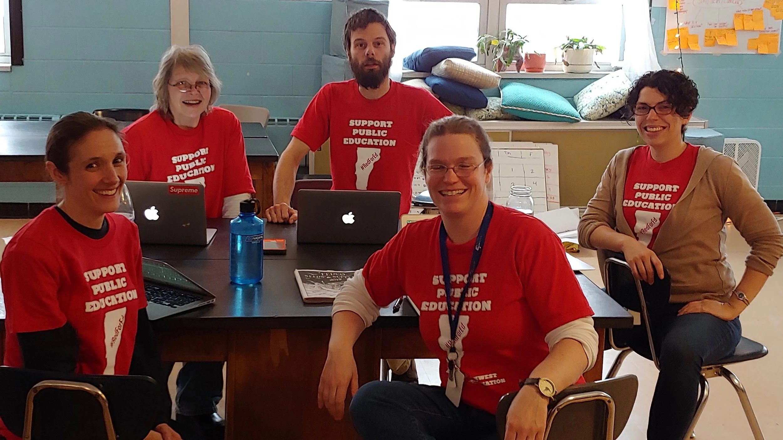 A group of six people in a room, all wearing red shirts with white text that reads 'Support Public Education.' They are sitting around a table with laptops, notebooks, and water bottles, and appear to be engaged in a meeting or discussion. There are 