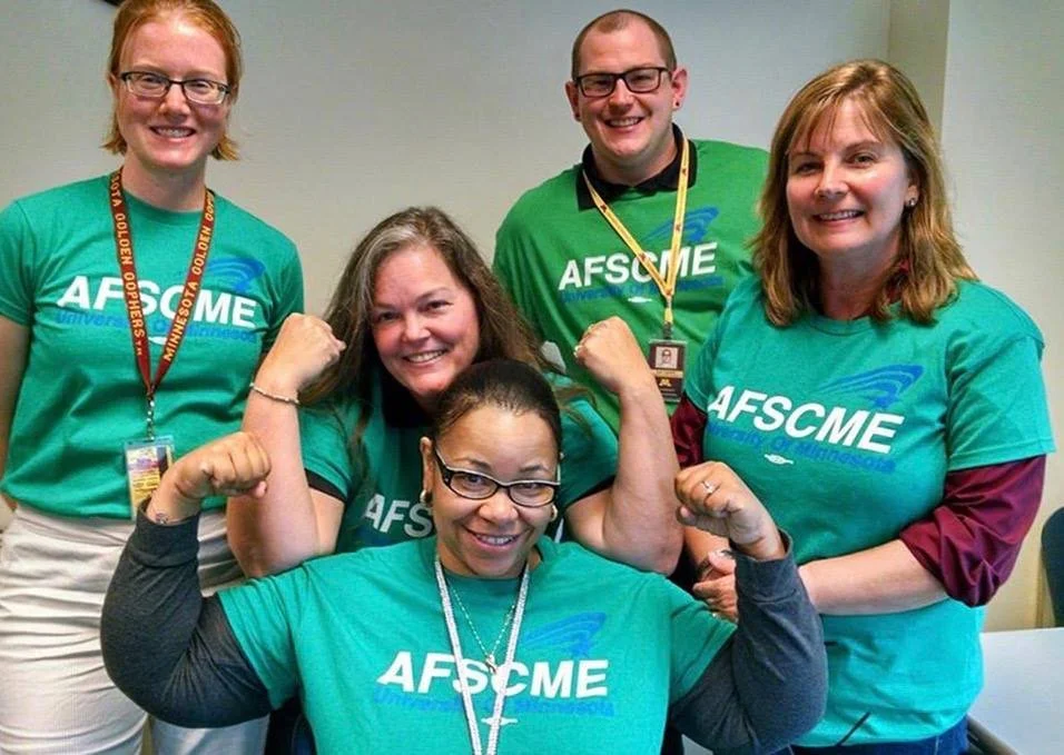 Group of five smiling people in teal AFSCME shirts, two women in front flexing muscles, one woman with glasses, one man and one woman with blonde hair, all smiling and showing team spirit.